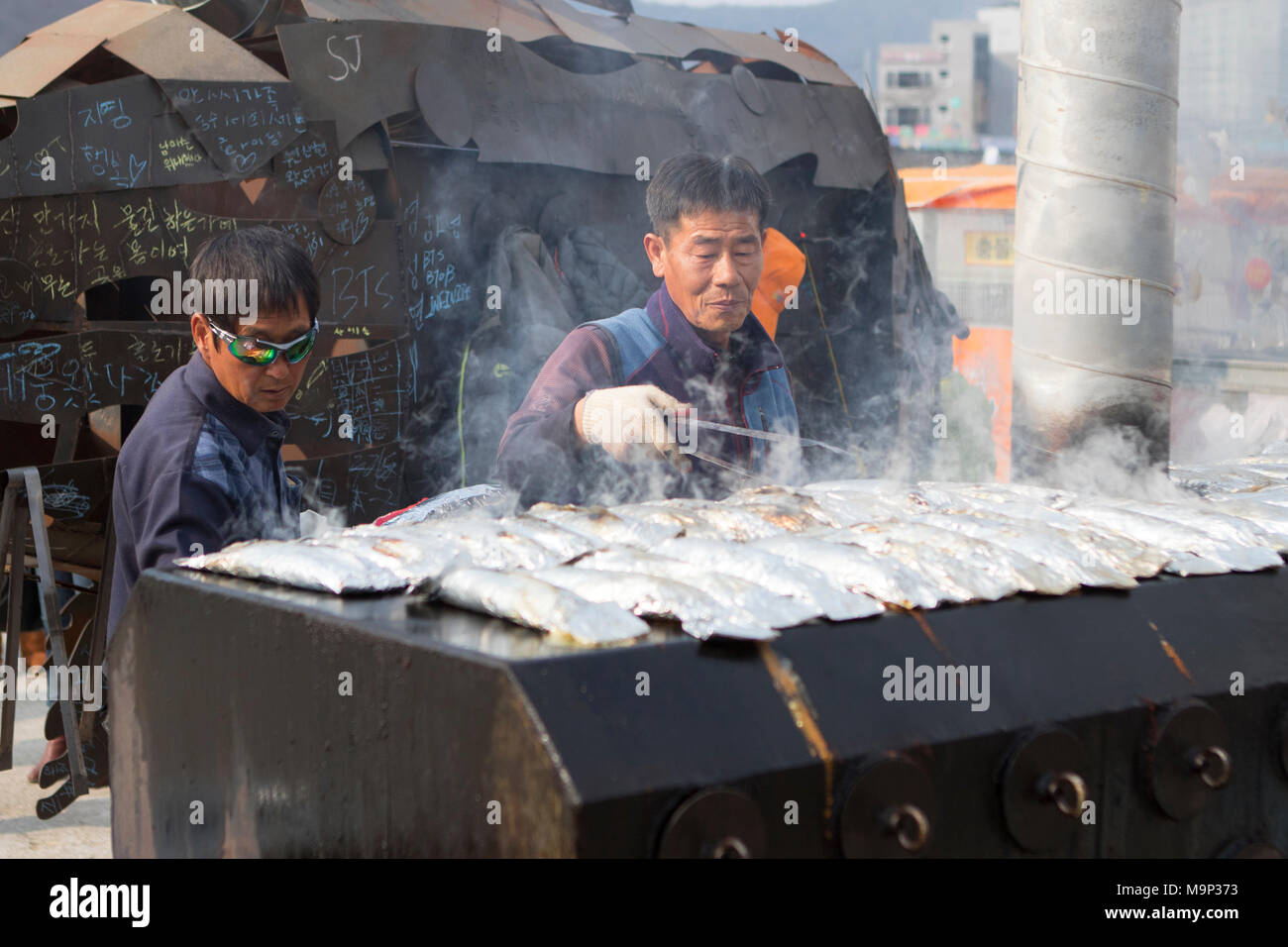 Un uomo è la cottura alla griglia il pesce appena pescato al Hwacheon Sancheoneo festival di ghiaccio. Il Hwacheon Sancheoneo Festival di ghiaccio è una tradizione per il popolo coreano. Ogni anno nel mese di gennaio la folla si riuniranno presso il fiume congelato per celebrare il freddo e la neve dell'inverno. Attrazione principale è la pesca sul ghiaccio. Giovani e vecchi attendere pazientemente su un piccolo buco nel ghiaccio per una trota di mordere. In tende possono lasciare il pesce grigliato dopo che loro sono mangiati. Tra le altre attività sono corse in slittino e pattinaggio sul ghiaccio. La vicina regione Pyeongchang ospiterà le Olimpiadi Invernali nel febbraio 2018. Foto Stock