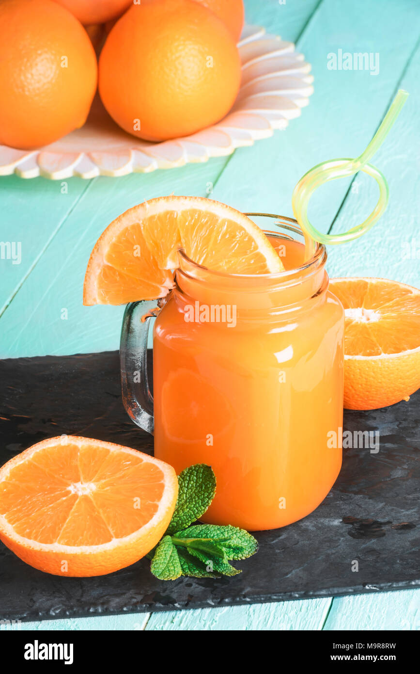 Mason vaso pieno di succo d'arancia fresco, con un divertente di paglia, circondato da fette di arance e le foglie di menta e una piastra di arance in thr sfondo. Foto Stock