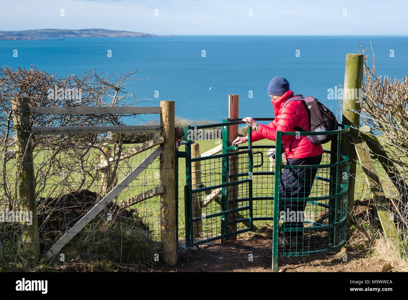 Anziani attivi donna senior walker a piedi attraverso un nuovo metallo kissing cancello vicino costa con mare oltre. Llanddona Isola di Anglesey Wales UK Gran Bretagna Foto Stock