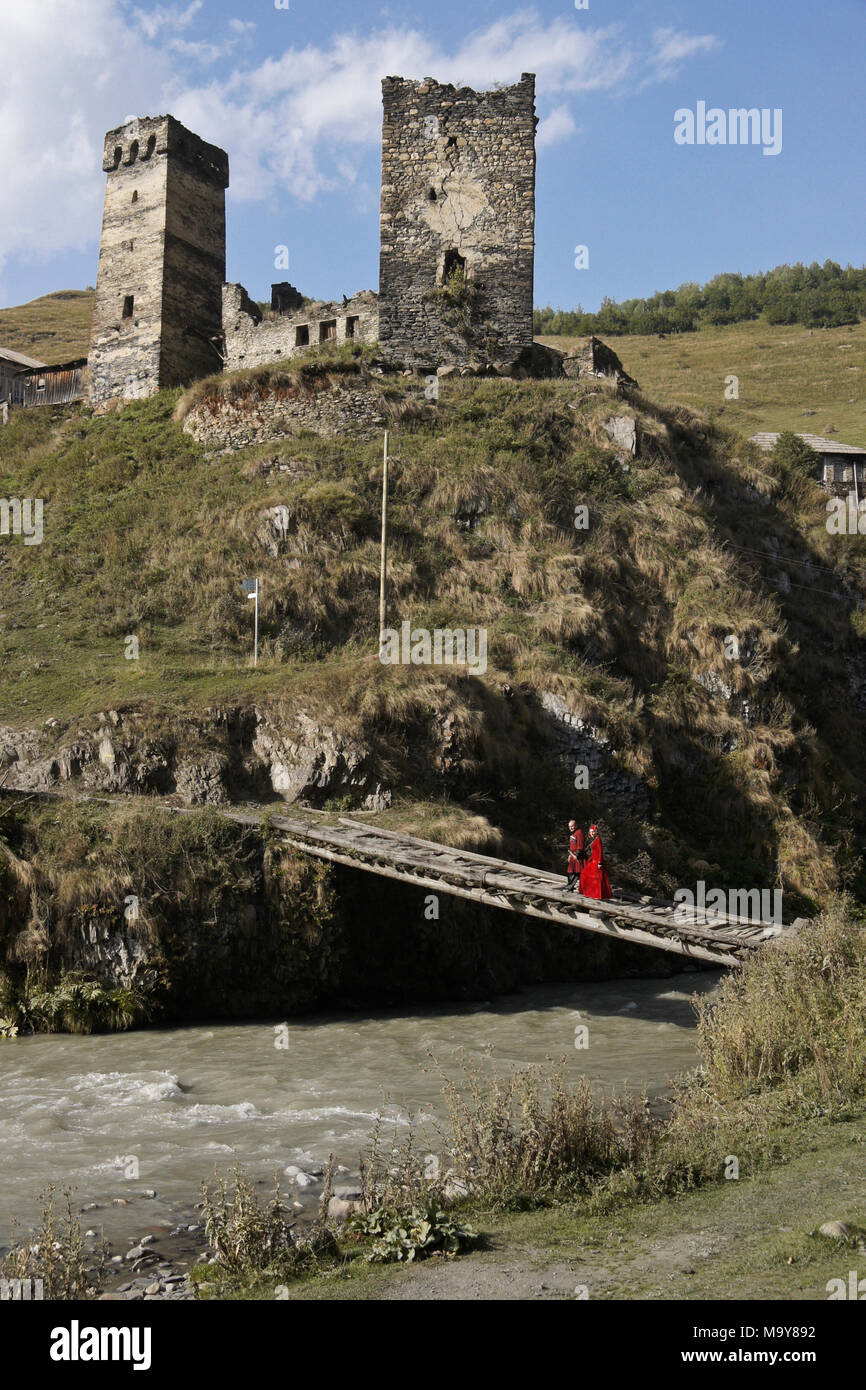 Un paio in Georgian National dress attraversare un ponte di legno sopra l'Inguri (Enguri) fiume sottostante medievale torri in pietra, Svaneti superiore, Georgia Foto Stock