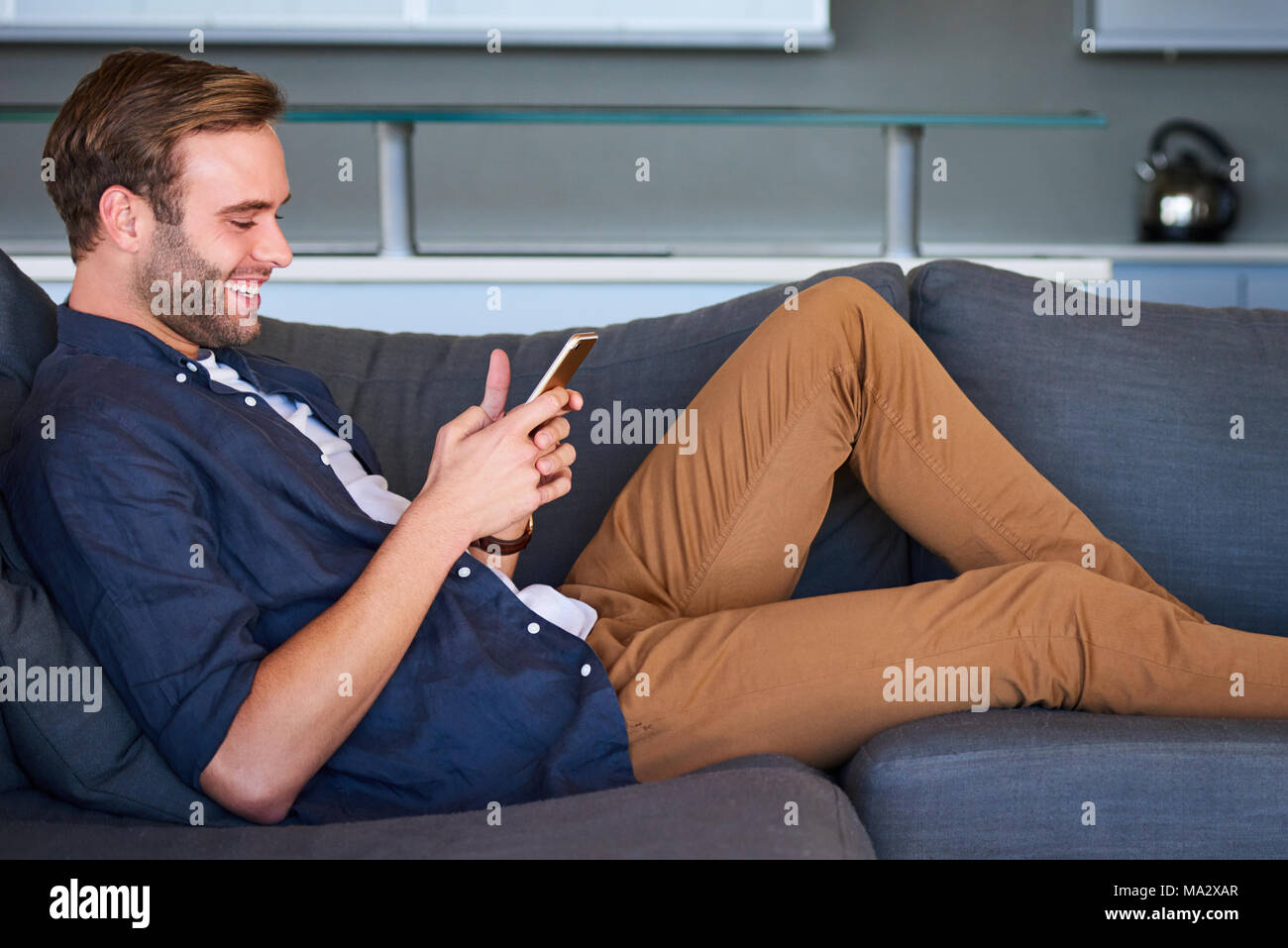 Immagine del profilo di uomo bello occupato sorridere mentre gli sms sul suo telefono cellulare, comodamente seduto sul suo lettino moderno nel suo salotto con il design elegante Foto Stock