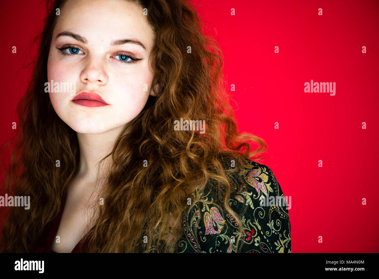 In prossimità della faccia di un moody affascinante giovane 13 tredici anni teenage ragazza adolescente con lunghi capelli rossi contro uno sfondo di colore rosso, in una foto studio , REGNO UNITO Foto Stock