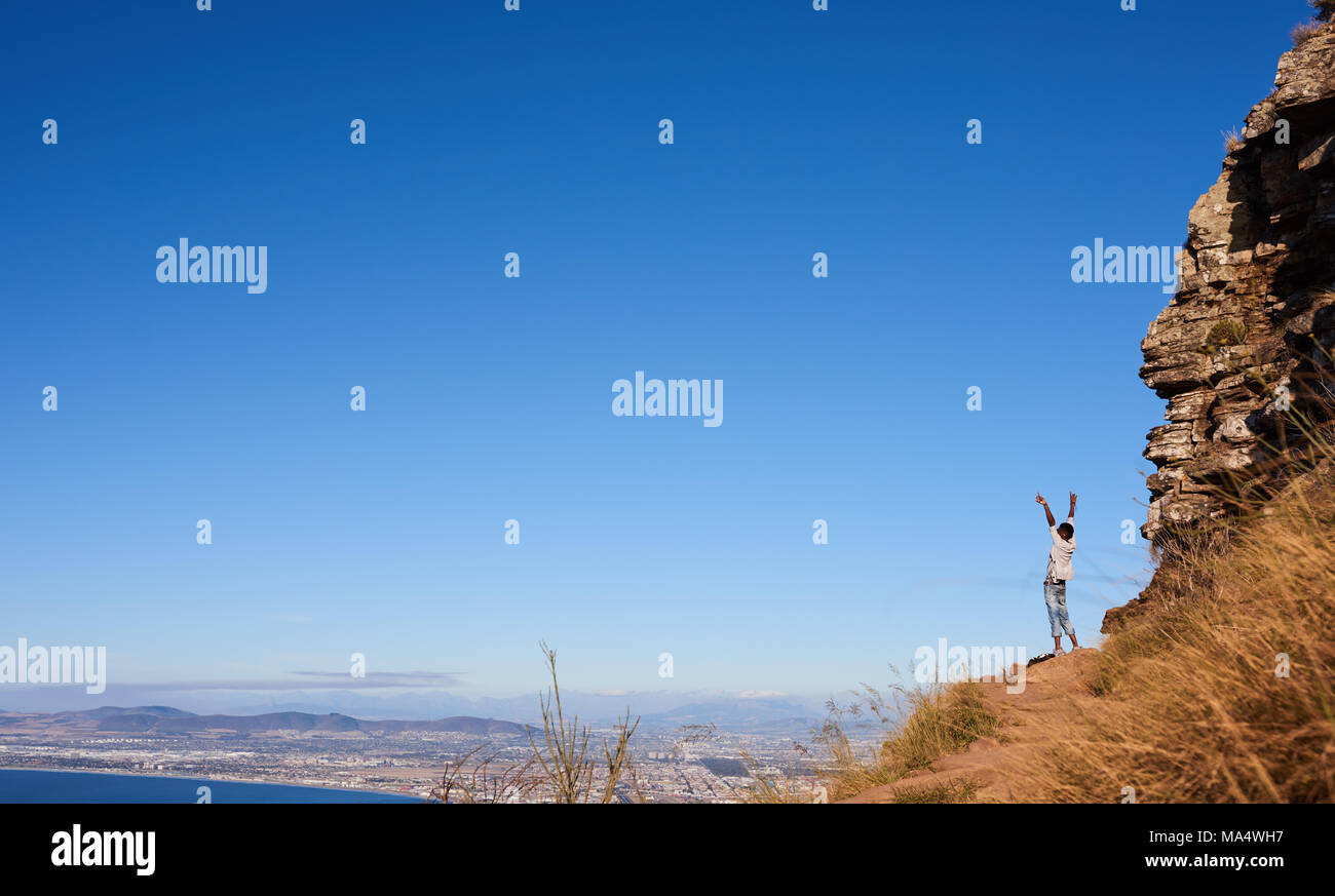 African uomo in piedi su una montagna all'aperto con le braccia e le mani aperte di fronte ad una vista magnifica e cielo blu chiaro su una città incredibile scape. Foto Stock