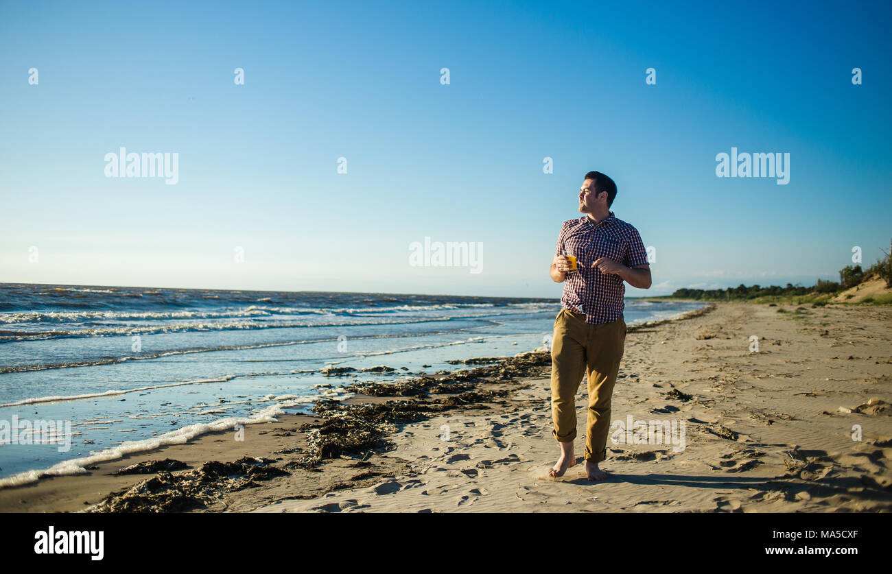 Felice uomo bello sorridere di fronte all'oceano con un bicchiere di succo di agrumi, indossare comode abiti leggeri. Foto Stock