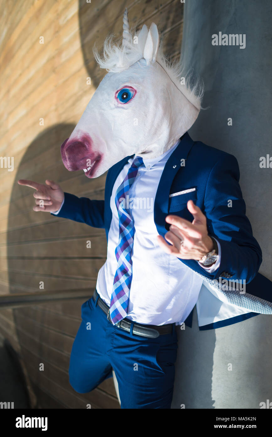 Giovani manager di successo in abito elegante e divertente maschera gesticulate attivo. Guy insoliti punti per voi. Unicorn diffonde le mani e rilassante sul banco di lavoro Foto Stock
