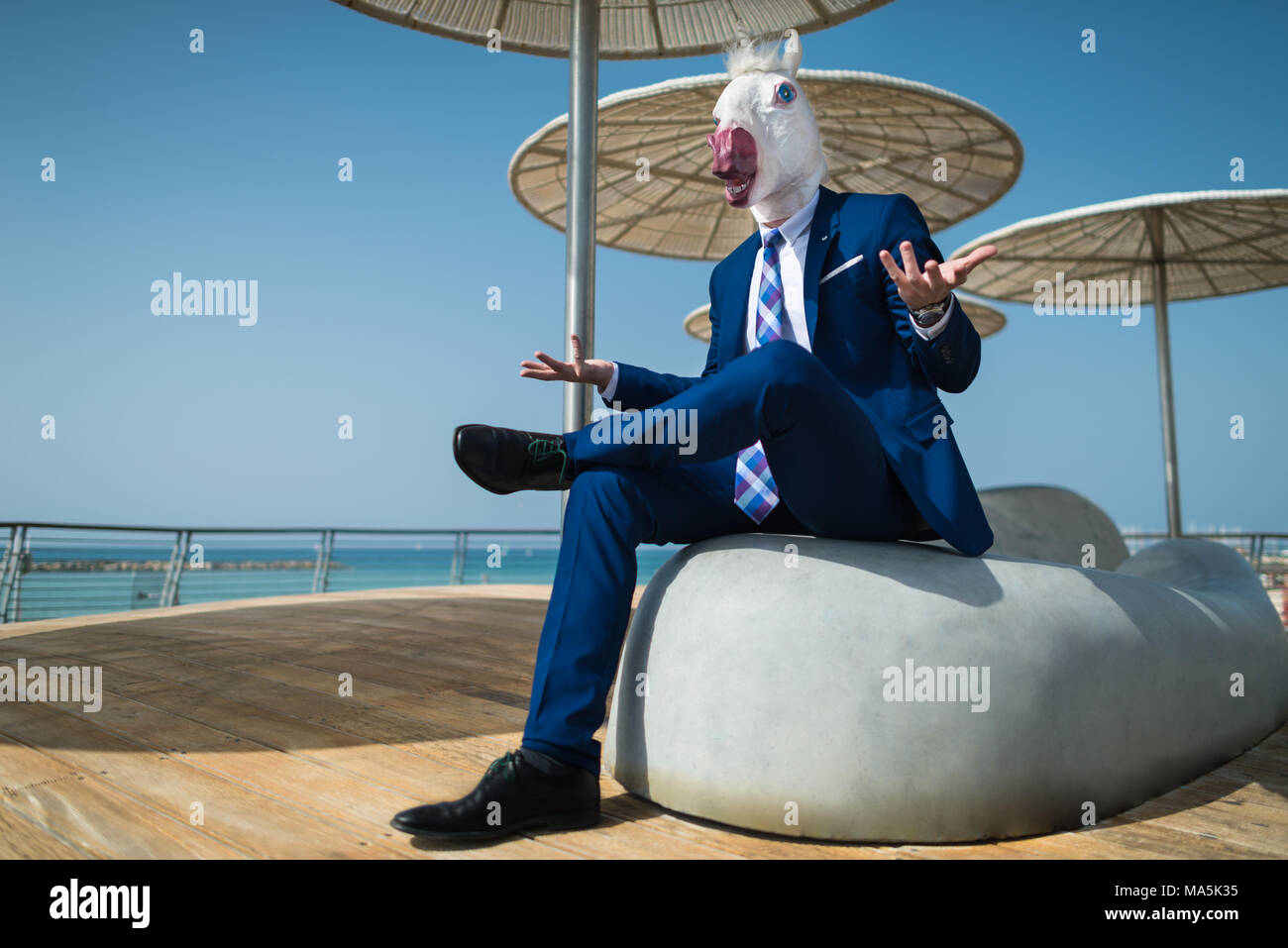 Giovane uomo in tuta siede sotto gli ombrelloni sul lungomare della città e gesticulate attivo. Manager di insolito nella maschera di comico si diffonde le mani. Elegante unicorn Foto Stock