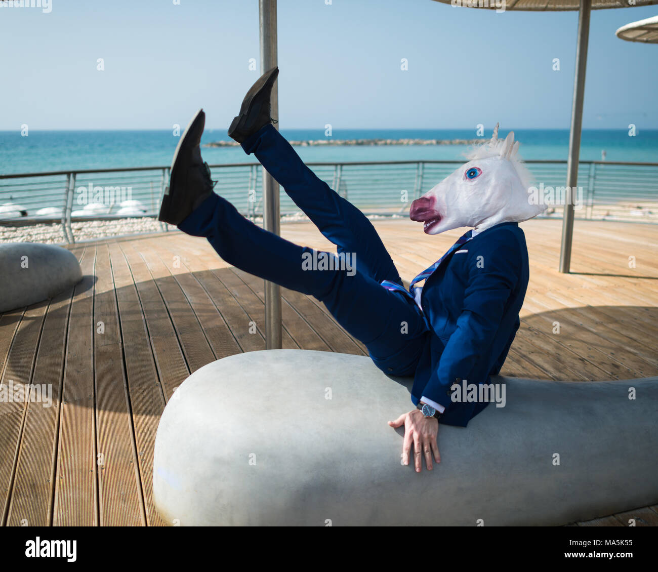 Giovane uomo in tuta scende sotto gli ombrelloni sul lungomare della città. Imprenditore inusuale in divertente maschera alla passeggiata del lungomare. Unicorn in posa comico Foto Stock