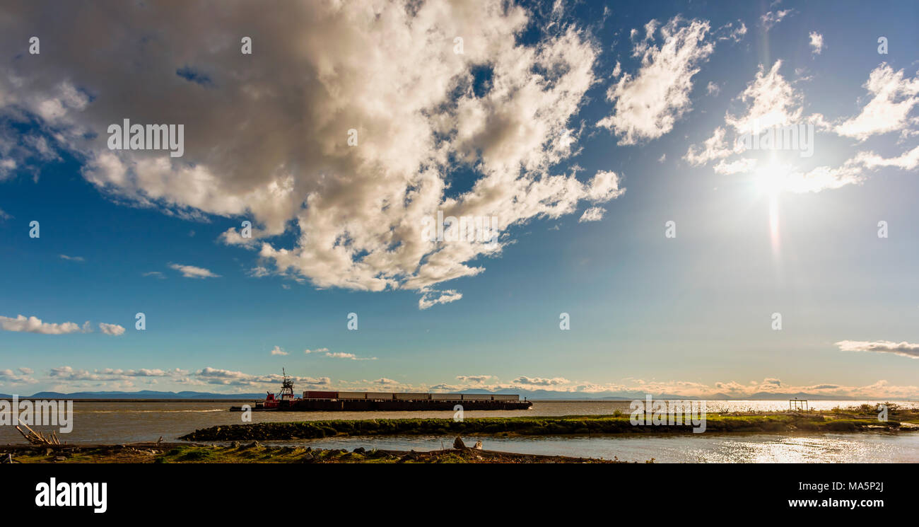 Una vista dalla banca ricoperte di erba e pietre sul fiume con una barca spingendo la chiatta sull'acqua e montagne sullo sfondo e un enorme s Foto Stock