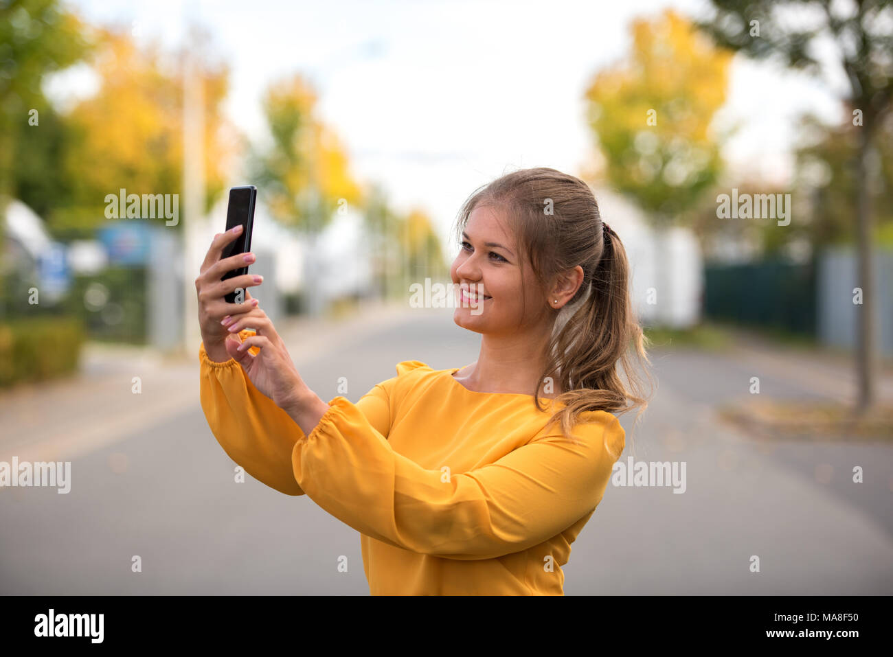 Giovane donna felice indossando abito giallo tenendo selfie con lo smartphone sulla strada Foto Stock Giovane donna felice indossando abito giallo tenendo selfie con lo smartphone sulla strada Foto Stock