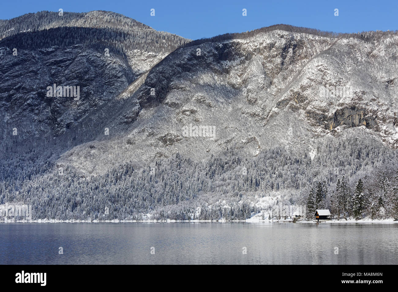 Piccola cabina in corrispondenza del piede di un lago con boschi innevati e montagne sullo sfondo Foto Stock