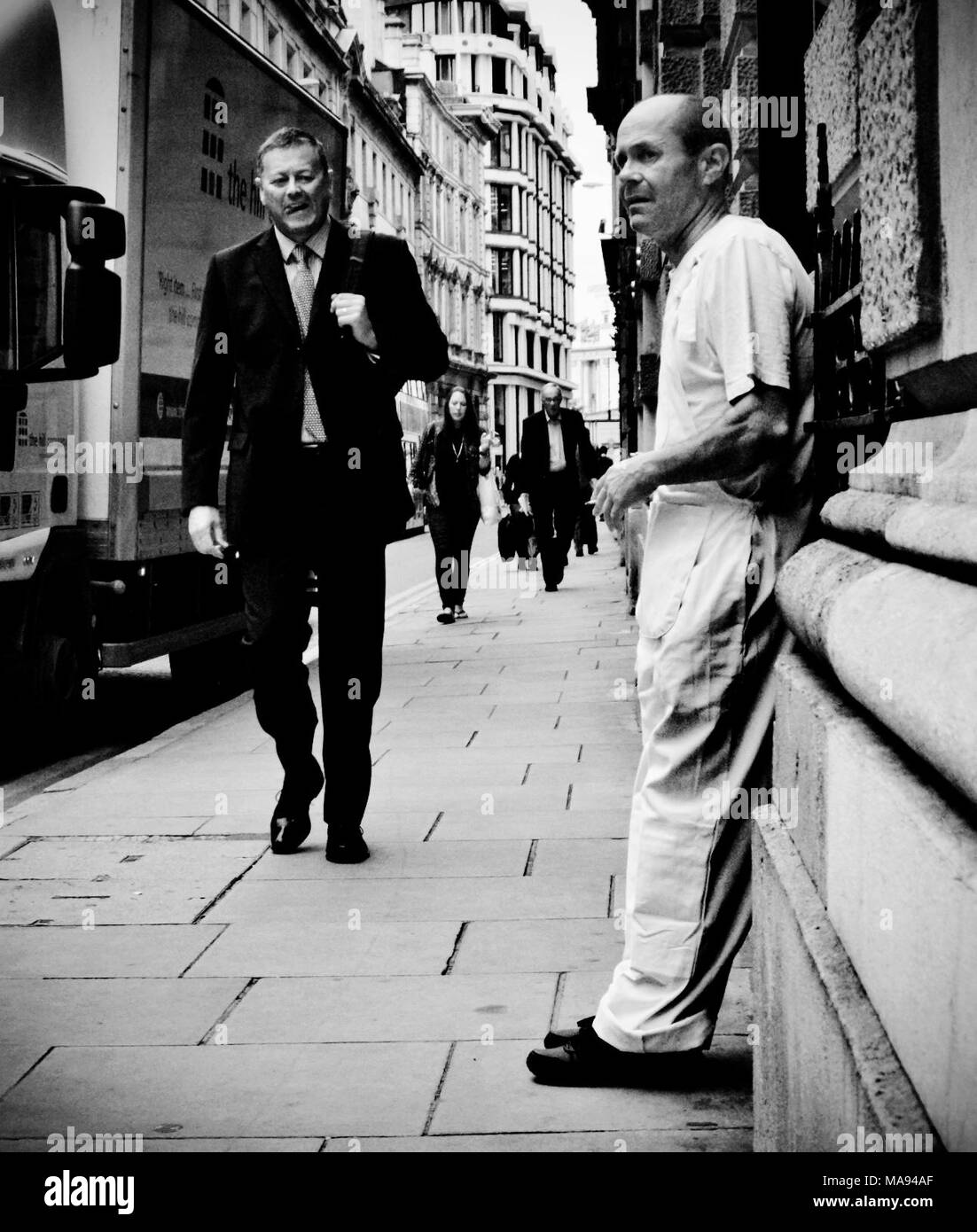Un tuttofare prende una pausa sigaretta come un lavoratore di ufficio passeggiate passato lui nella città di Londra. Foto Stock