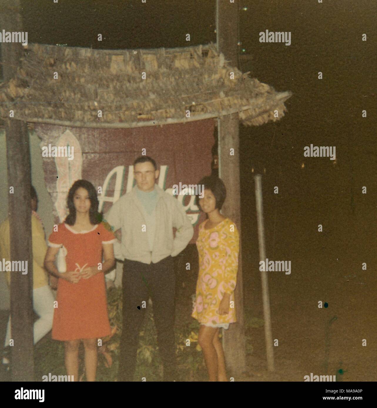 Fotografia a colori che mostra un uomo in quasi pieno-view, indossa una giacca grigia, in piedi di fronte a un segno, fiancheggiata da due donne, sia indossando abiti colorati, un'altra persona sta dietro un segno pilastro a sinistra, fotografato in Vietnam durante la Guerra del Vietnam (1955-1975), 1968. () Foto Stock
