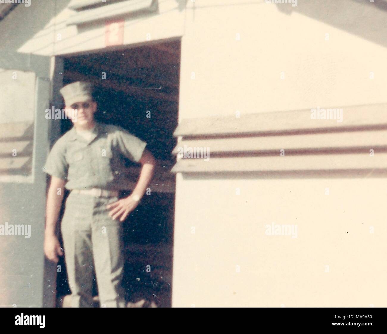 Fotografia a colori, girato da un basso angolo di un soldato di sesso maschile, in vista in tre quarti, che indossa una uniforme, occhiali da sole e cappello, rivolto verso la fotocamera con un espressione rilassata sul suo volto, mentre in piedi in una porta aperta, fotografato in Vietnam durante la Guerra del Vietnam, 1968. () Foto Stock