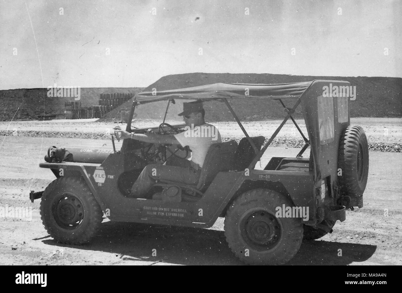 Fotografia in bianco e nero che mostra un maschio di Marine, seduto dietro al volante di una jeep contrassegnata USMC, parcheggiato su una strada sterrata in un paesaggio roccioso e regolazione del lato conducente specchio retrovisore, con pallet di forniture impilati nella parte anteriore di una collina sullo sfondo, fotografato in Vietnam durante la Guerra del Vietnam (1955-1975), 1968. () Foto Stock