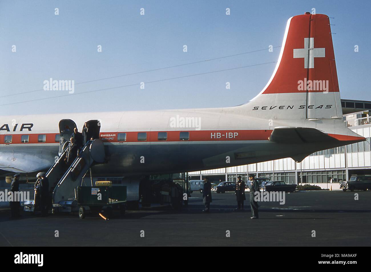 Vista della sezione di coda della fusoliera di un Swissair DC 7C HB IBP 'Sanche dei mari" aeromobili parcheggiati fuori della international terminal arrivi a Edoardo Vianello New York International Airport, Giamaica, Queens, a New York City, Giugno 1959. Passeggeri uscire dal piano porta giù per una scalinata direttamente sull'asfalto a bordo sinistro. I dipendenti della compagnia aerea salutare le persone lo sbarco, come New York City poliziotti guardare in basso a destra. () Foto Stock