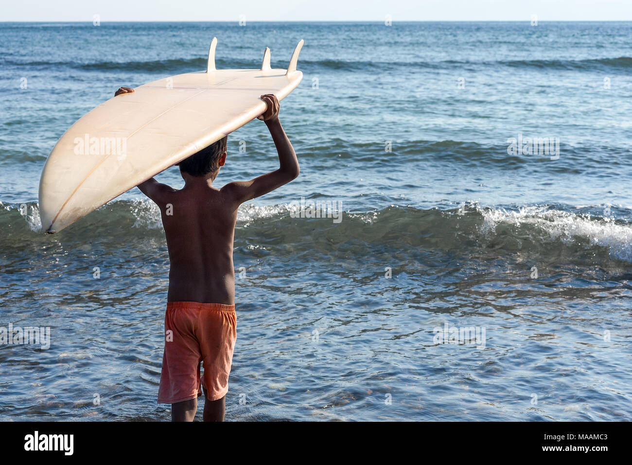 Un locale giovane ragazzo indonesiano in pantaloncini usurato porta il suo donato tavole da surf sulla sua testa in un mare calmo. Foto Stock