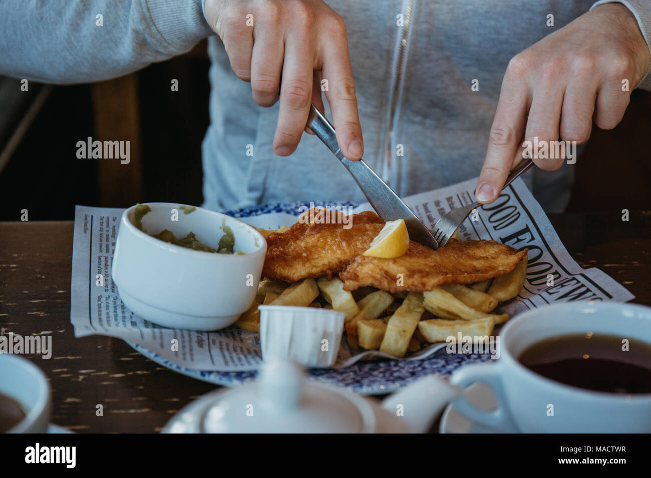 Uomo seduto al tavolo di legno, mangiando pesce e patatine, Inglese tradizionale piatto, tè e salse vicino a. Foto Stock