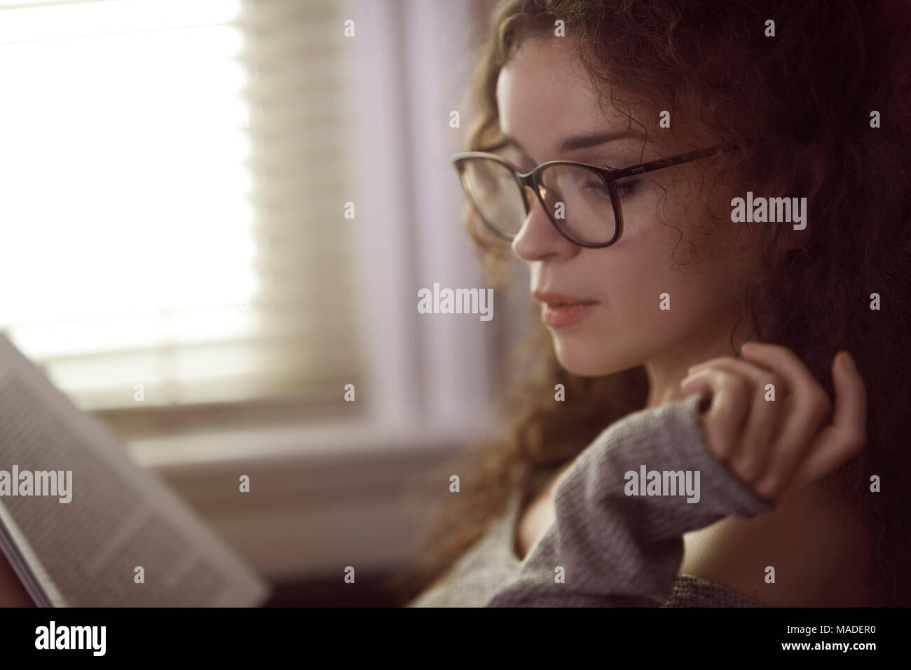 Ritratto di una giovane donna con gli occhiali per leggere un libro dalla finestra nella luce solare Foto Stock