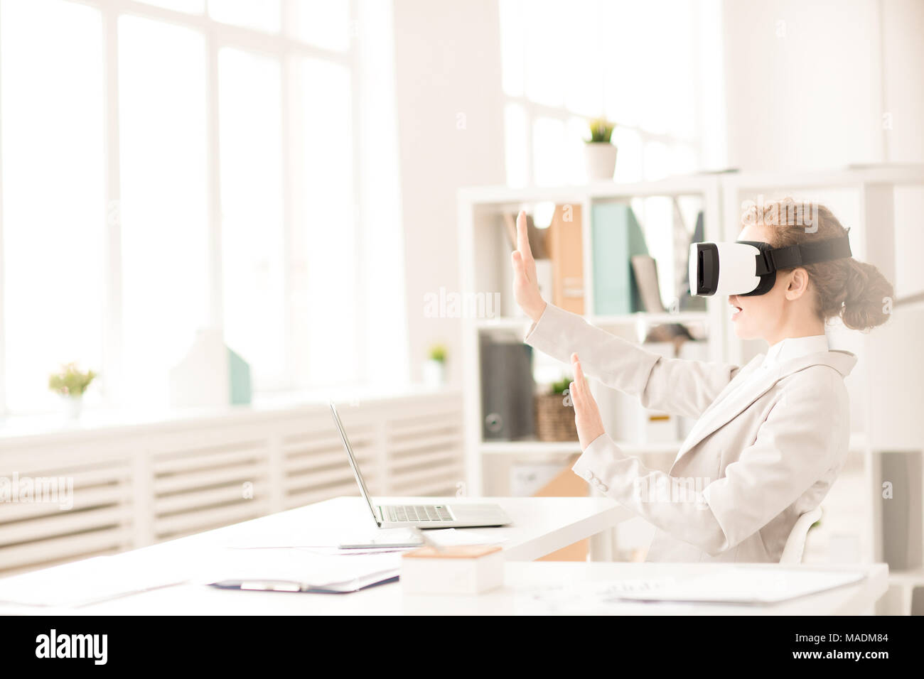 Imprenditrice indossando occhiali per realtà virtuale per il computer portatile e gesticolando seduto alla scrivania in ufficio Foto Stock
