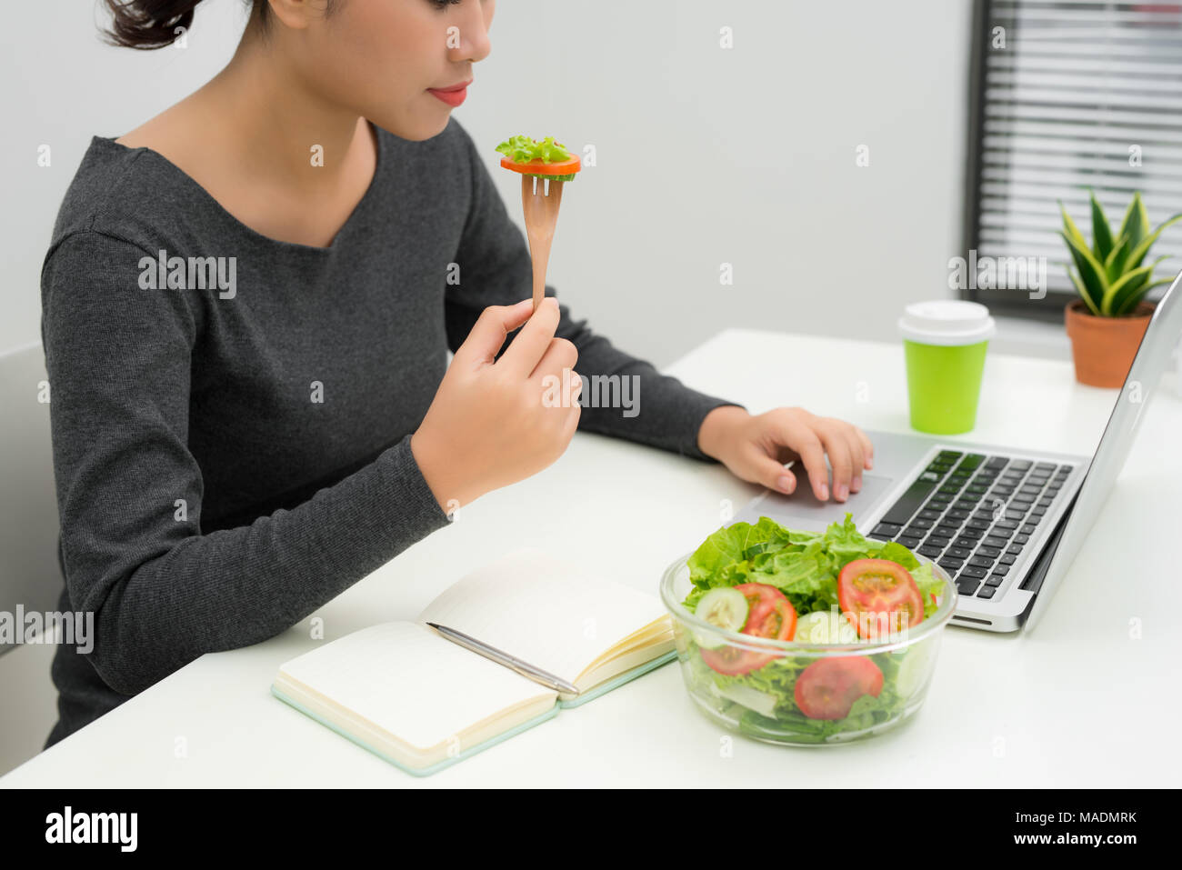 La donna ha un sano pranzo di lavoro in un ufficio moderno interno. Bella giovane imprenditrice al posto di lavoro, mangiare insalata di verdure nella ciotola, dieta e v Foto Stock