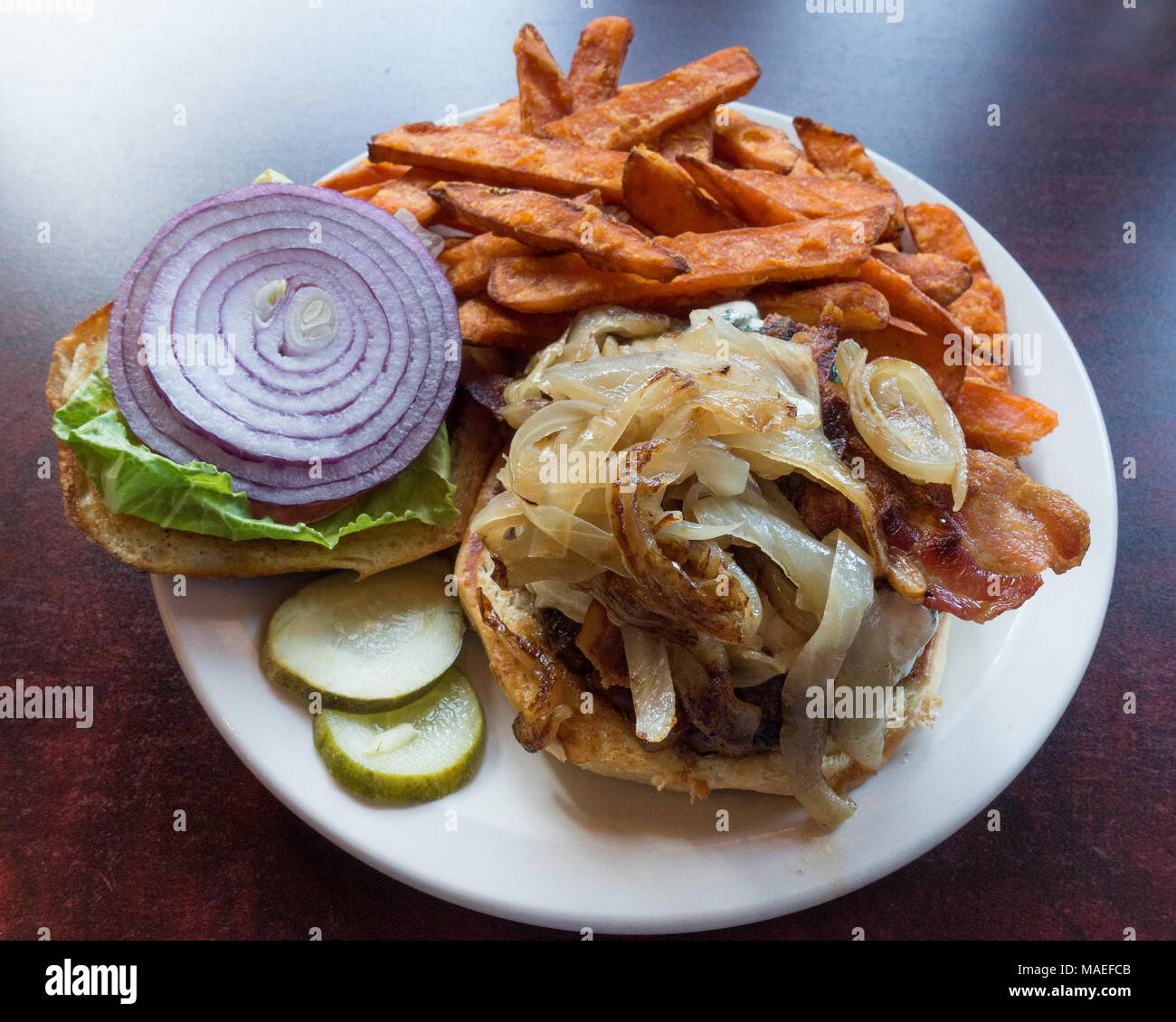 Un gourmet cheeseburger con formaggio blu, pancetta e cipolle su un rullo morbido servito con lattuga, cipolla e pomodoro, sottaceti e patate dolci patatine fritte. Foto Stock