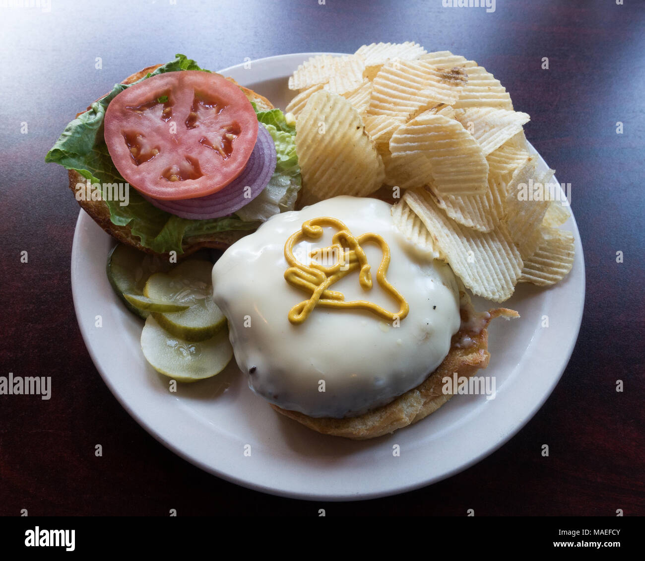 Un gourmet cheeseburger con lattuga, cipolla e pomodoro su un rullo morbido servito su una piastra bianca con patate fritte e sottaceti. Foto Stock
