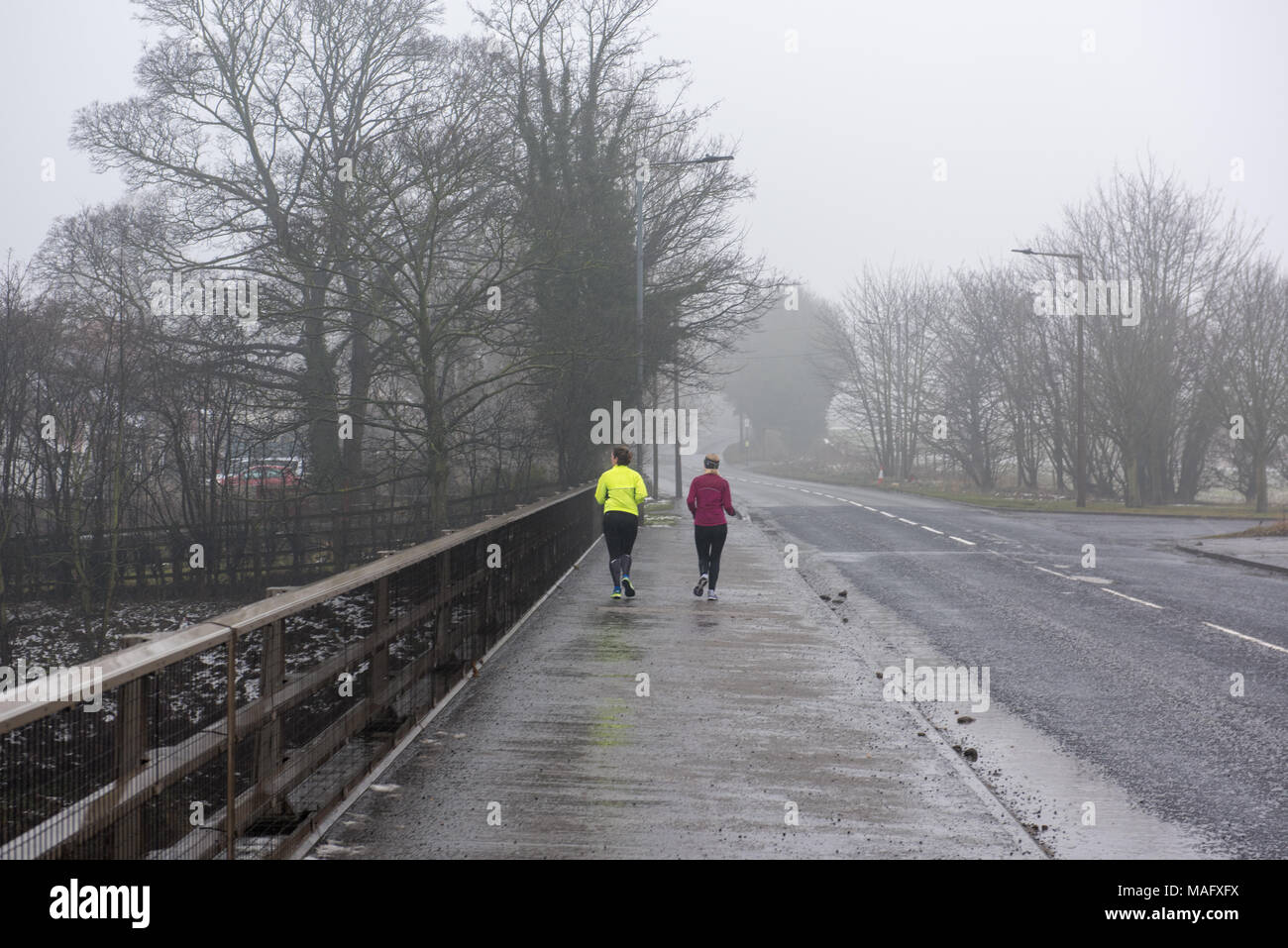 Due jogger che corrono lungo una strada nebbiosa in una nebbiosa mattina invernale a Doncaster, South Yorkshire, Inghilterra Foto Stock