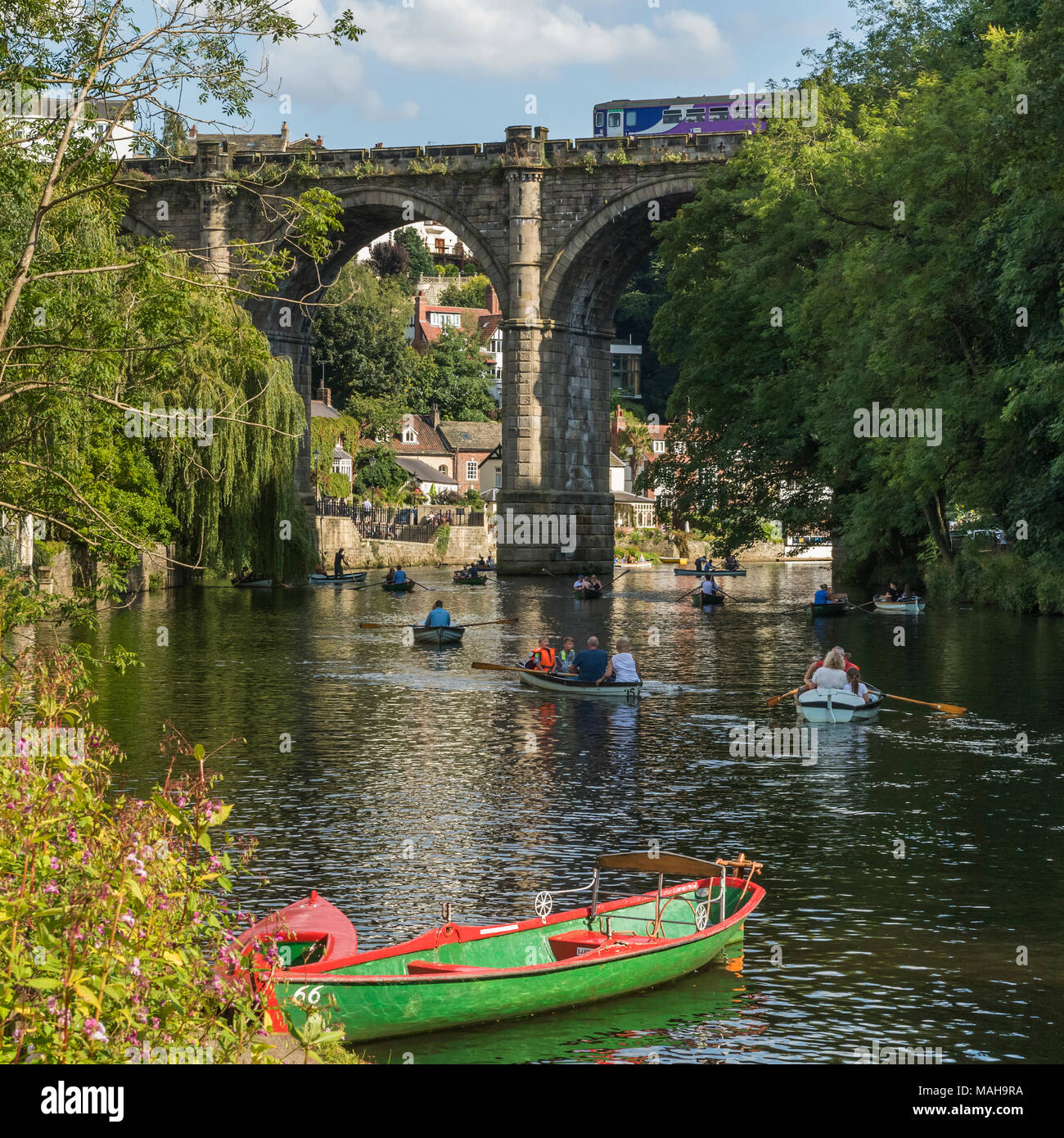 Persone e rilassanti gite in barca a remi sul fiume Nidd sotto il cielo estivo blu, come il treno passa sul ponte in scenic sunny Knaresborough, England, Regno Unito Foto Stock
