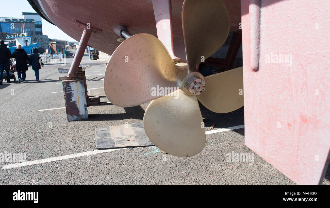 Primo piano di un'elica di una barca in ottone su un'imbarcazione nel bacino di carenaggio in un porto turistico, con persone che camminano sullo sfondo, in Inghilterra Foto Stock