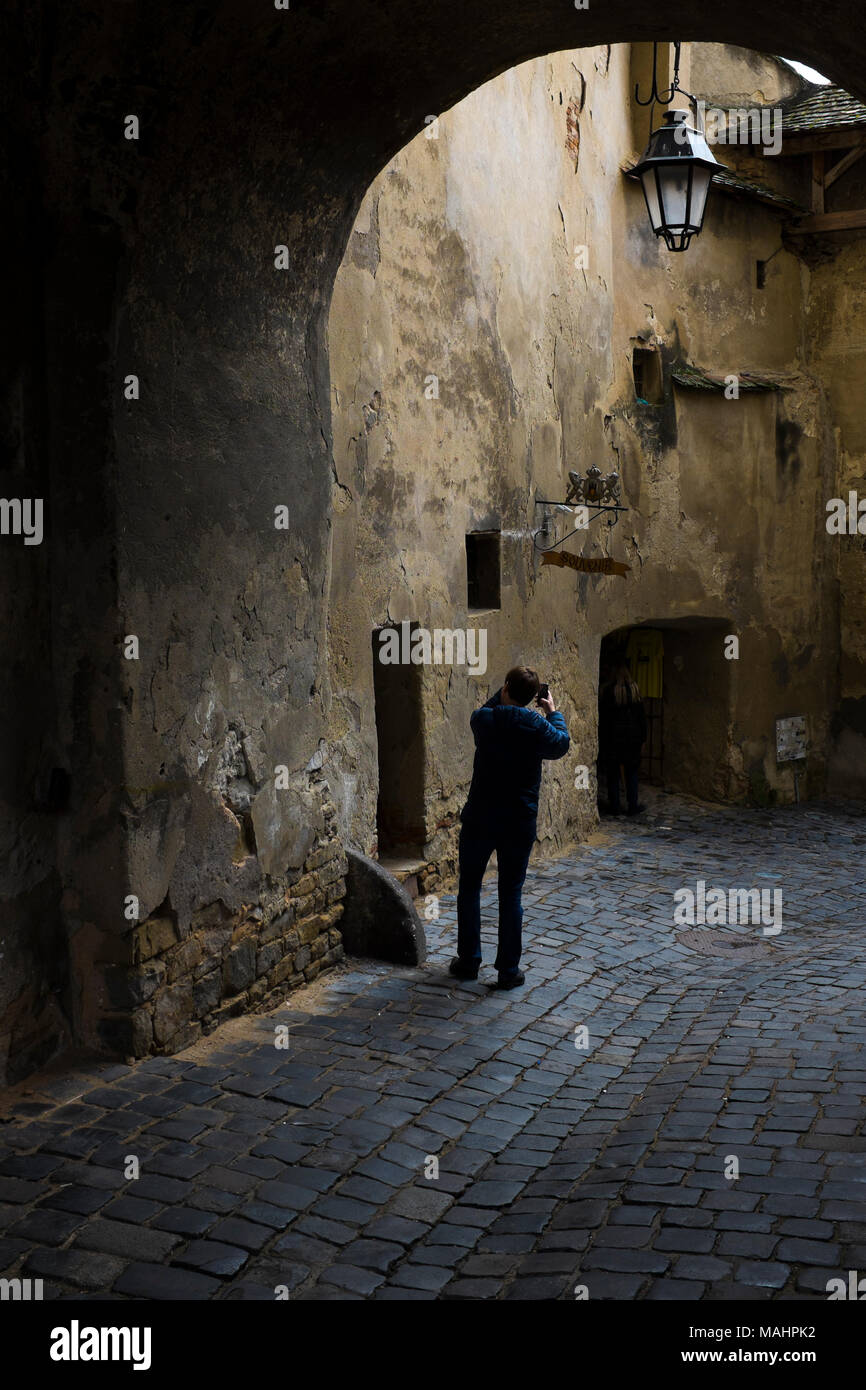 Un uomo prende una fotografia sul lastricato, strette e colorate strade avvolgimento fino alla cittadella nella città vecchia di Sighisoara, Transilvania, Romania. Foto Stock