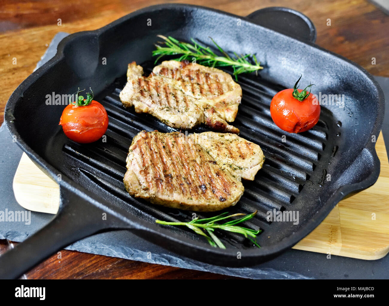 Le bistecche grigliate in una padella di ferro con pomodori e il ramo di rosmarino. Bistecche grigliate su un tavolo in legno, cucina gourmet. Foto Stock