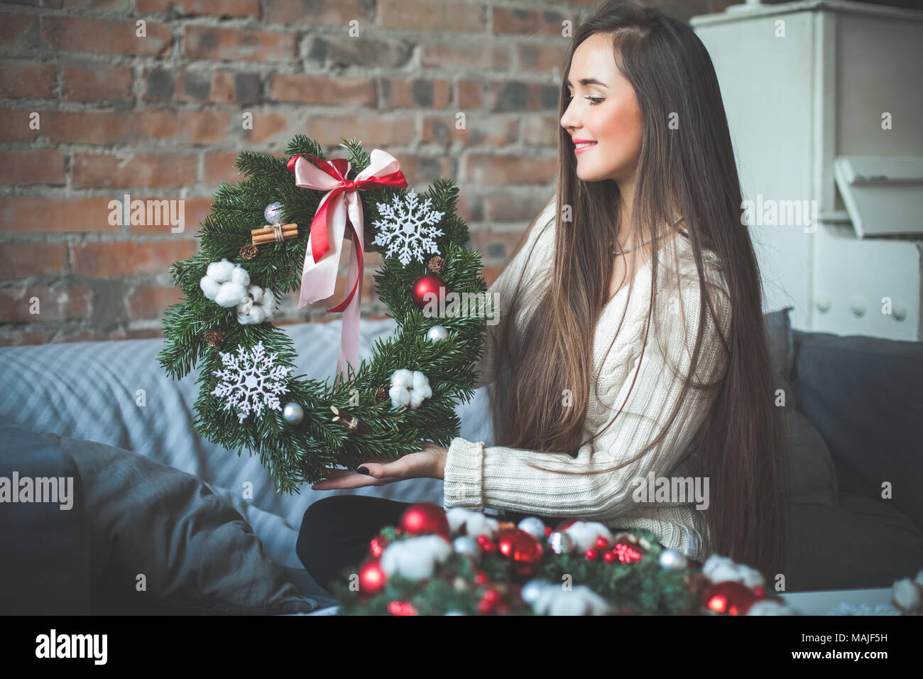 Felice fioraio di giovane donna seduta a casa sul luogo di lavoro e far Natale albero sempreverde Ghirlanda rosso con sfere di vetro, coni, nastro e cannella. Vero Foto Stock