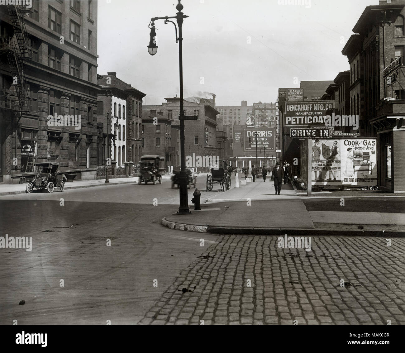 In orizzontale e di fotografia in bianco e nero di una street view del angolo di Broadway e Cerre strade. Carrozze trainate da cavalli e automobili viaggiano lungo la sporcizia strada lastricata che è rivestita con mattoni e pietra di edifici commerciali. Annunci per 'Denckhoff Mfg. Co Manufacturing Sottovesti', 'Drive nell', 'Dgiaggiolo camion', ' San Louis Poster Adv. Co.', 'McKnight Sartoria Co.', 'Cook-Gas Detroit gioiello Hellrung & Grimm' e 'YWCA'. Titolo: Guardando ad est su Locust Street (Luogo Lucas) dalla sedicesima Street. . 1914. Swekosky, William G., 1895-1964 Foto Stock