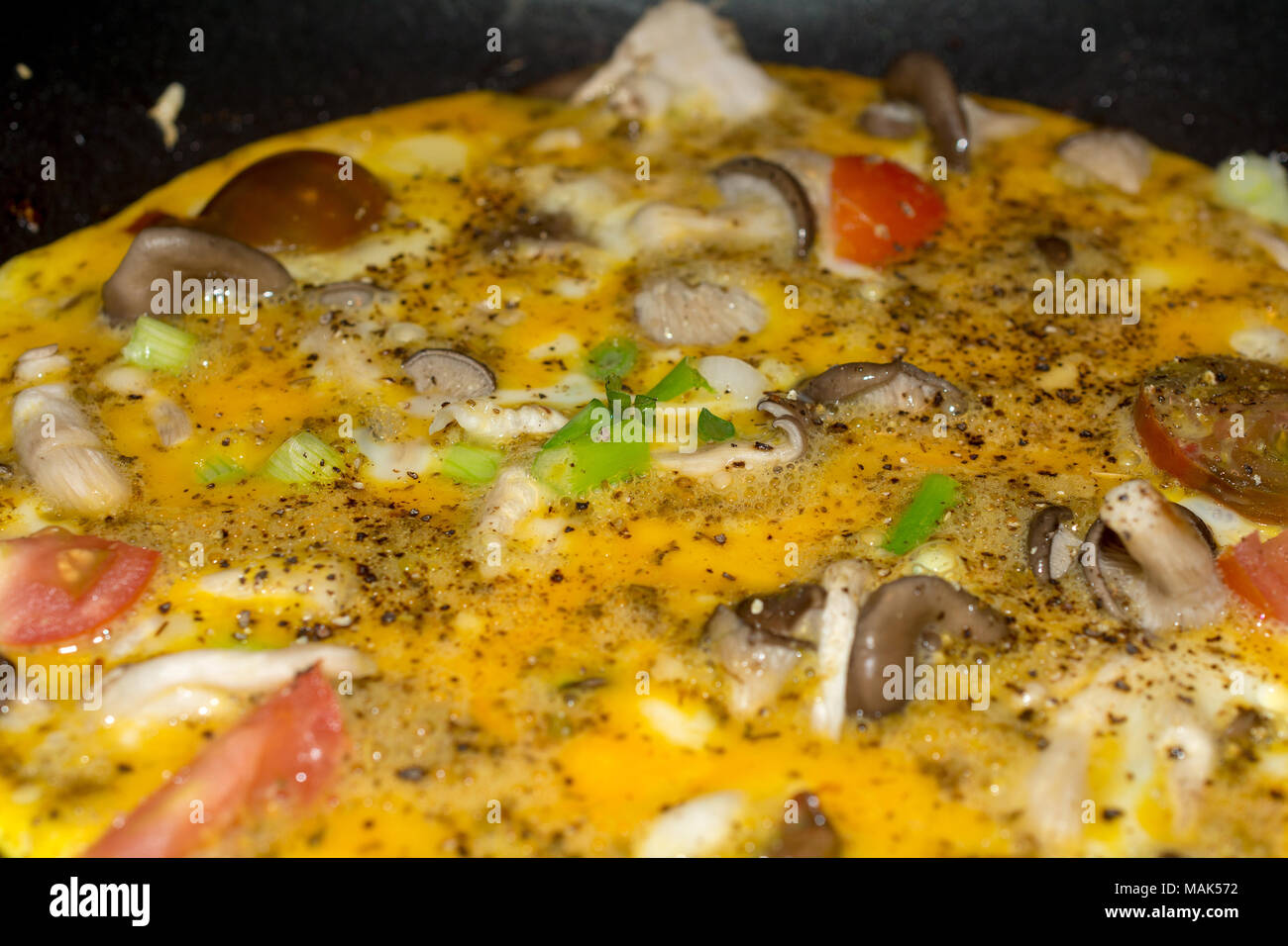 Cucinare una frittata di close-up omelette cucinare in padella in ghisa con funghi, pomodori e cipolla a molla e guarnita con pepe nero Foto Stock