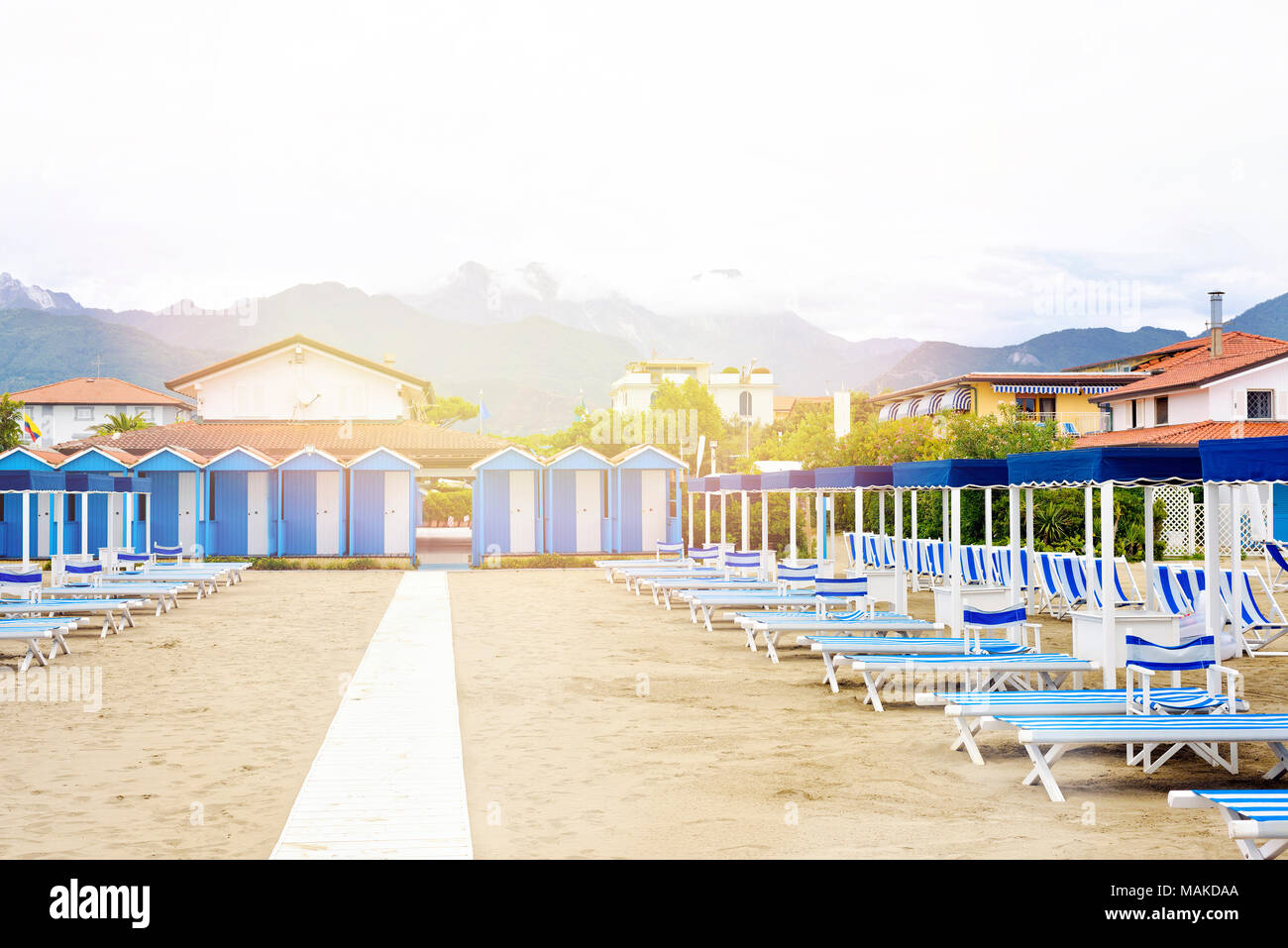 Vista diurna alla vibrante blu sdraie e ombrelloni sulla spiaggia. Cielo nuvoloso, edifici e montagne sullo sfondo. Copia negativo spazio, luogo per il testo Foto Stock