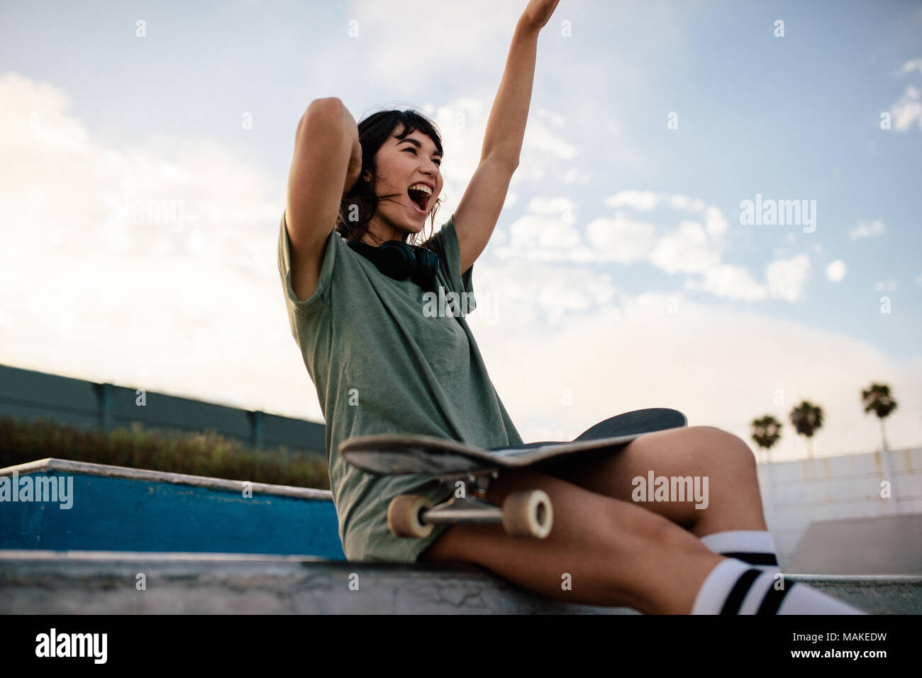 Urbano sorridente ragazza seduta sulla rampa da skateboard in skate park avendo divertimento. Guidatore di skateboard femmina che si gode all'aperto a skate park. Foto Stock