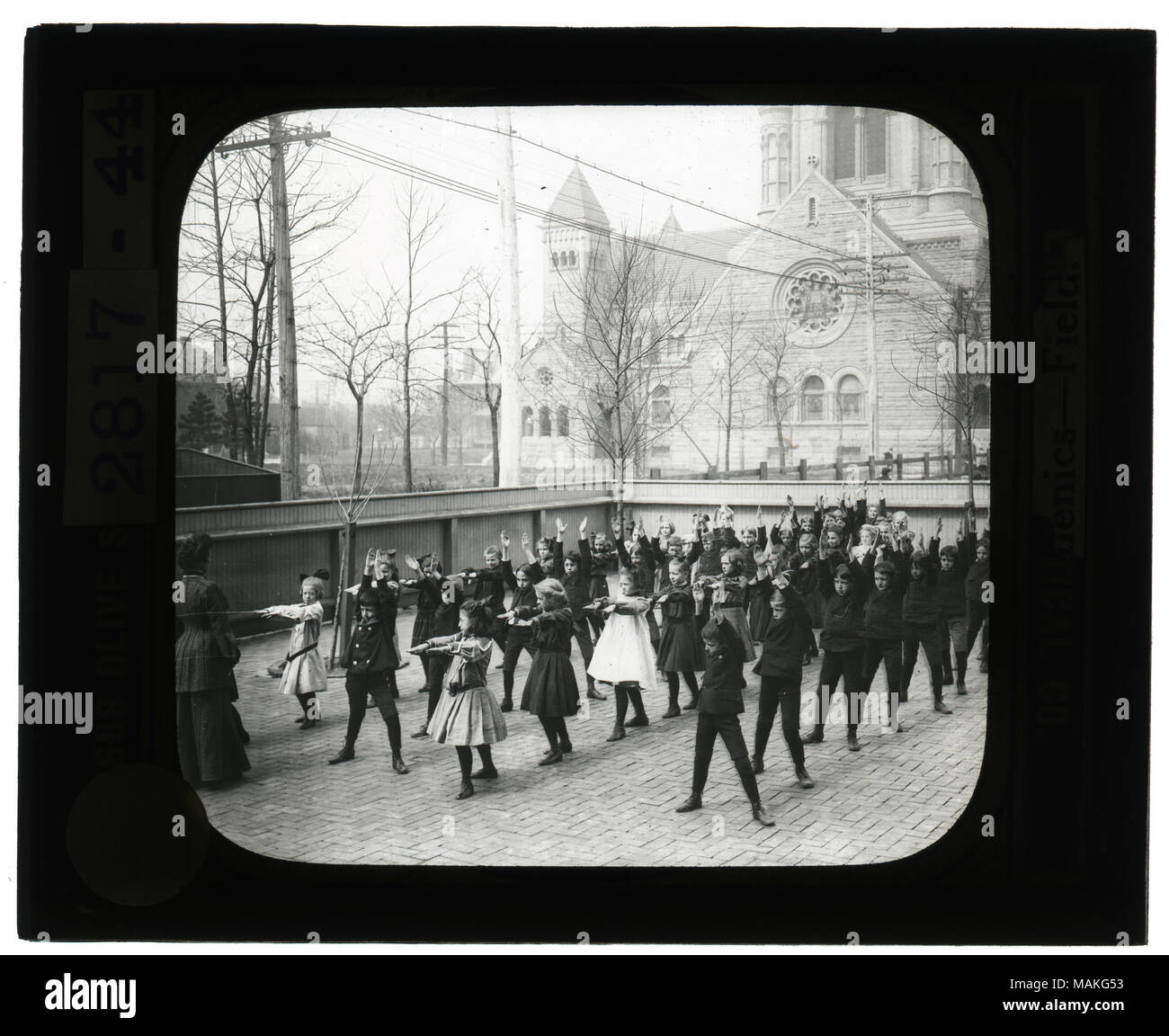 Orizzontale, foto in bianco e nero di ragazzi e ragazze al di fuori su di un mattone cortile pavimentato facendo calisthenics. Il cantiere dispone di una recinzione di legno intorno ad esso e non vi è una grande chiesa in background. Titolo: museo educativo lantern slide: Calisthenics- campo scuola. . Circa 1904. Foto Stock Orizzontale, foto in bianco e nero di ragazzi e ragazze al di fuori su di un mattone cortile pavimentato facendo calisthenics. Il cantiere dispone di una recinzione di legno intorno ad esso e non vi è una grande chiesa in background. Titolo: museo educativo lantern slide: Calisthenics- campo scuola. . Circa 1904. Foto Stock