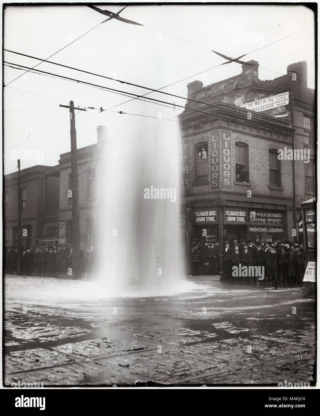 Verticale, fotografia in bianco e nero che mostra una folla guarda il pennacchio da rottura di un idrante di fuoco al di fuori dei fratelli Fellhauer saloon e negozio di liquori a 900 Market Street. Il geyser dall'idrante di fuoco è vicino al centro dell'immagine e la folla è imballato sul marciapiedi dietro di esso. I due piani di un edificio di mattoni di alloggiamento dei fratelli Fellhauer negozio di liquori è sull'angolo di una strada a destra, con insegne pubblicitarie liquori medicinali, whiskey e vini. Le linee di alimentazione di criss-cross attraverso la metà superiore dell'immagine. Vedere il retro della stampa per brevi note relative a indirizzi di Fe Foto Stock