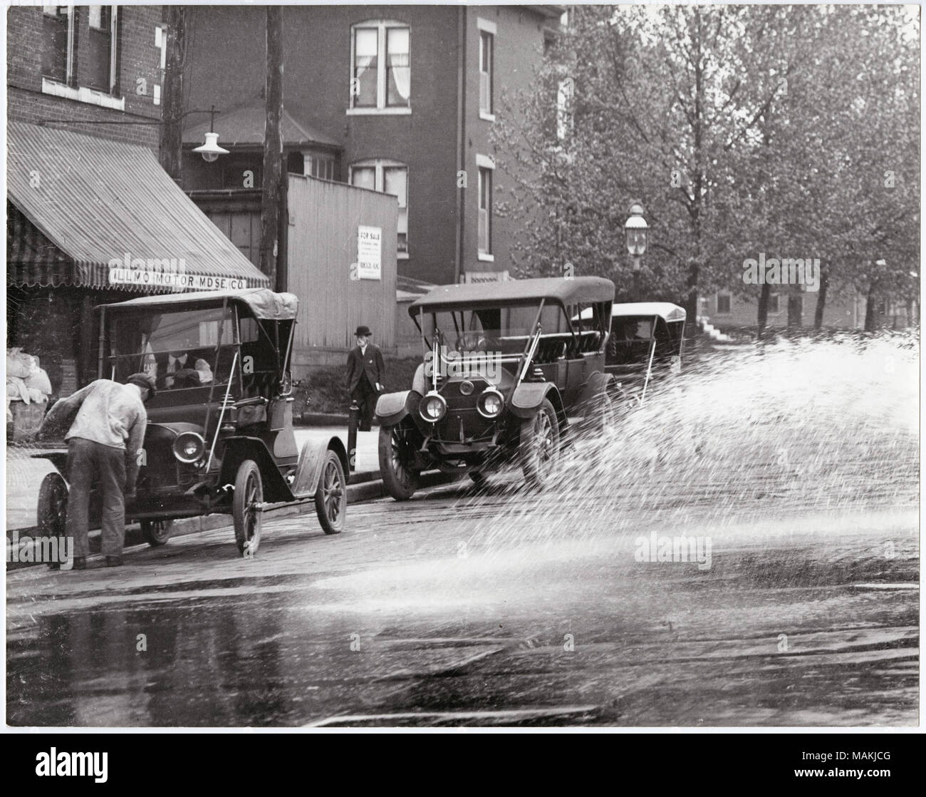 In orizzontale e di fotografia in bianco e nero che mostra tre 1910-era l'auto parcheggiata di fronte al motore Illmo Merchandise Company. Il conducente è seduto nella prima vettura, mentre un altro uomo utilizza una mano-manovella per avviare il motore. Un autista è visibile anche nell'ultima vettura. Un grande spruzzo di acqua, probabilmente da una strada carrello sprinkler, germogli nel lato destro dell'immagine. Titolo: automobili parcheggiate fuori il motorino Illmo Merchandise Company. . Circa 1910. Holt, Charles Clemente, 1866-1925 Foto Stock