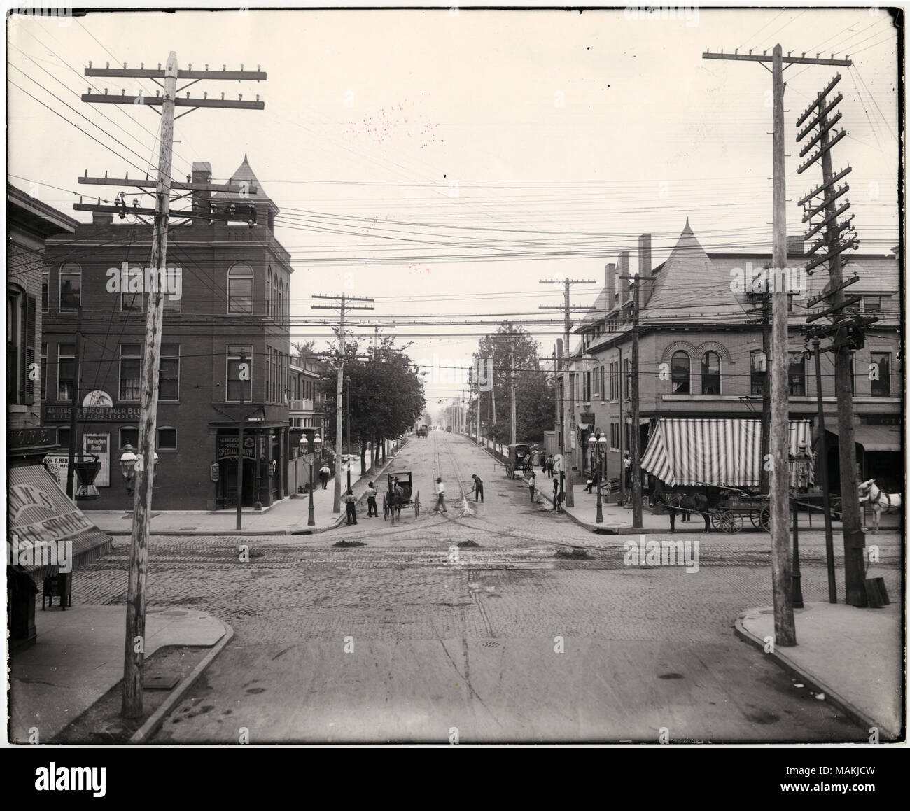 In orizzontale e di fotografia in bianco e nero che mostra la costruzione di edifici commerciali in corrispondenza dell'intersezione di Marcus e Easton, ora il dottor Martin Luther King Drive. La foto è stata scattata guardando verso il basso Marcus e comprende 4651 Easton sulla destra. Le imprese indicate includono anche una farmacia e un salone, eventualmente quella di Charles Bohn. Il salone è dotato di più pubblicità per Anheuser-Busch. Alcuni vagoni può essere visto in strada, tra cui una pubblicità della Dougherty fratelli business. Afro-americano di pedoni può essere visto sul marciapiede, e i lavoratori sono spazzare la strada. Titolo: edifici commerciali presso il Foto Stock