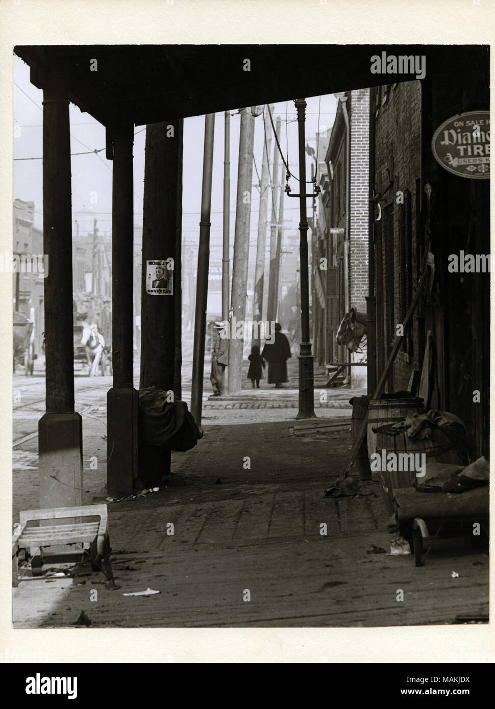 Verticale, fotografia in bianco e nero che mostra una madre e un bambino a camminare lungo il marciapiede di una strada non identificato. Supporto di pilastri di una tettoia che copre il marciapiede in primo piano. Di barili, pile di panni, e gli strumenti sono sparsi sotto la tettoia. Gli annunci pubblicitari possono essere visti per pneumatici di diamante e Michael J. Gill, un candidato per il Congresso. La costruzione di edifici commerciali la linea di entrambi i lati della strada, e carri trainati da cavalli possono essere visti sullo sfondo. Titolo: la madre e il bambino a camminare lungo il marciapiede su una strada non identificato. . Circa 1912. Holt, Charles Clemente, 1866-1925 Foto Stock