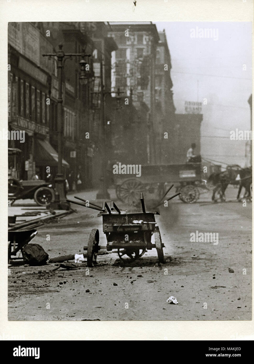 Verticale, fotografia in bianco e nero che mostra street riparare le attrezzature vicino all'angolo sud-ovest di Chestnut Street e 12th Street, ora Tucker Boulevard. Apparecchiatura include una carriola con le maniglie di vari strumenti da esso sporgente. Gli edifici commerciali di linea la strada a sinistra e una a cavallo il carro può essere visto in background. Titolo: Street attrezzature di riparazione nella strada vicino all'angolo sud-ovest di Chestnut Street e 12th Street, ora Tucker Boulevard. . Circa 1910. Holt, Charles Clemente, 1866-1925 Foto Stock