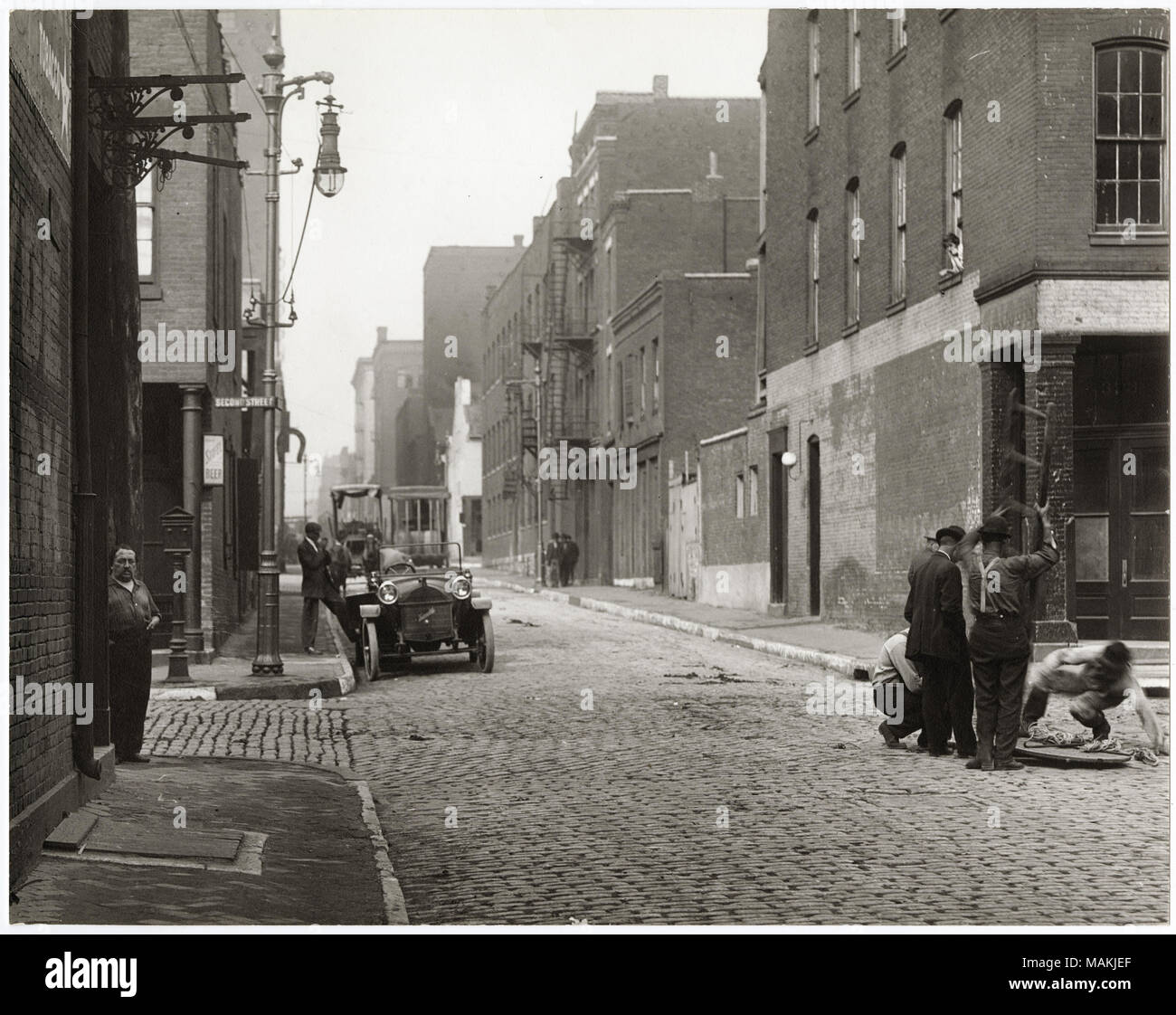 In orizzontale e di fotografia in bianco e nero che mostra un misterioso strada di ciottoli in corrispondenza di un angolo della seconda strada. Entrambi i lati della strada sono rivestiti con edifici in mattoni, variabile in altezza da due storie a quattro storie. Sul lato sinistro dell'immagine, un uomo è in piedi sull'angolo di strada sotto il cartello stradale per la seconda strada, mentre un altro uomo sorge accanto alla macchina parcheggiata sulla strada non identificato. Sul lato destro dell'immagine, un piccolo gruppo di uomini si sta abbassando una scaletta giù un tombino aperto. Nessun segni aziendali sono visibili, sebbene un segno per Stifel birra può essere visto al secondo angolo di strada. T Foto Stock