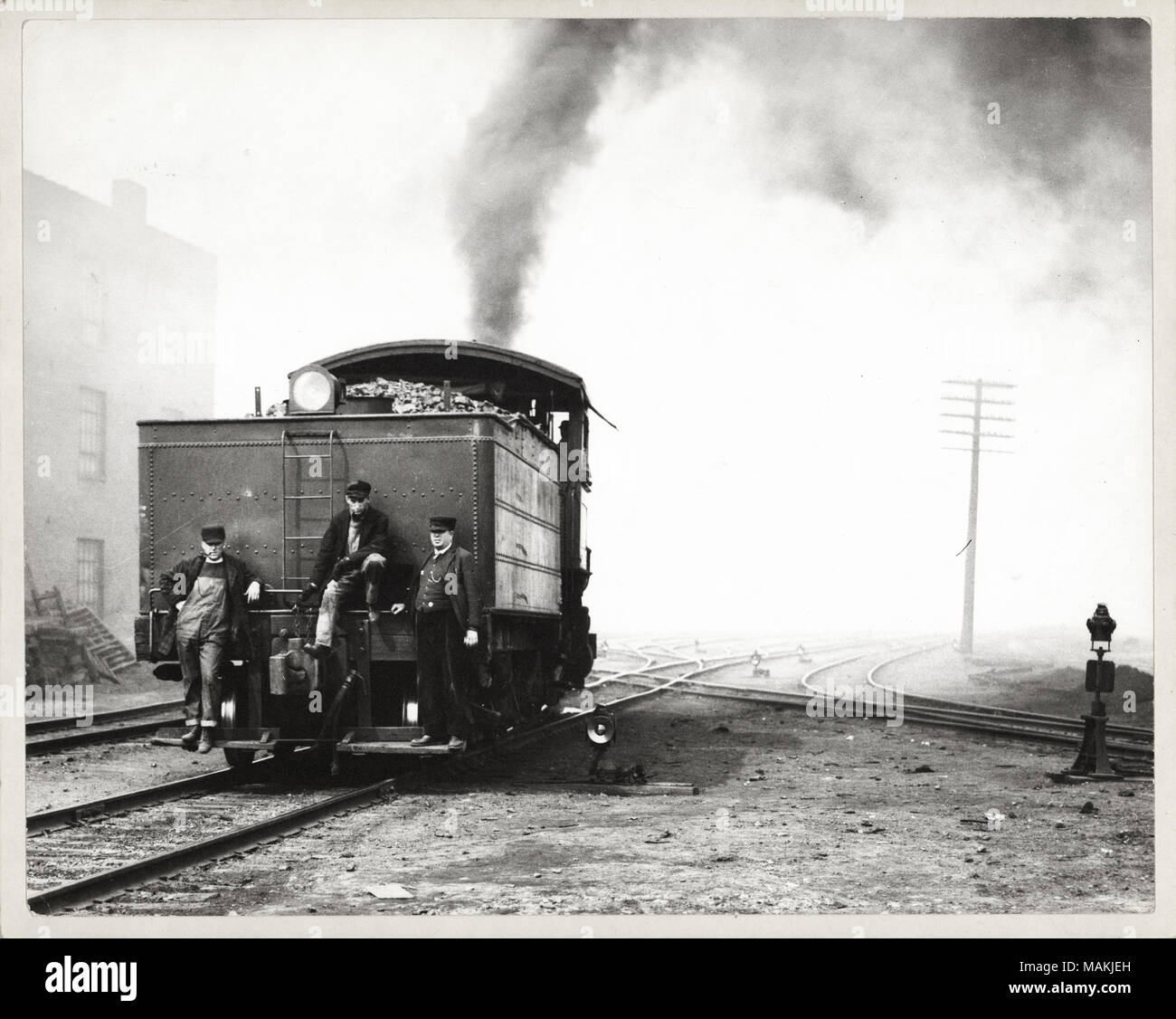 In orizzontale e di fotografia in bianco e nero che mostra tre uomini sul retro di un interruttore del vapore motore in un cantiere ferroviario. Due degli uomini stand su una scheda in esecuzione, mentre il terzo è arroccato su di una rotaia o tubazione. Un uomo può essere il conduttore, come egli indossa un cappello uniforme e una tuta, mentre gli altri due uomini usura operai di abbigliamento. Un faro è nella parte superiore di una scaletta corta e carbone accatastati nel bunker può essere visto dietro di esso. Un pennacchio di fumo flutti lontano dalla stazione. Diversi i binari della ferrovia criss-cross in background. Una nota manoscritta sul retro della stampa, probabilmente dalla Scuola Suore di Notre D Foto Stock