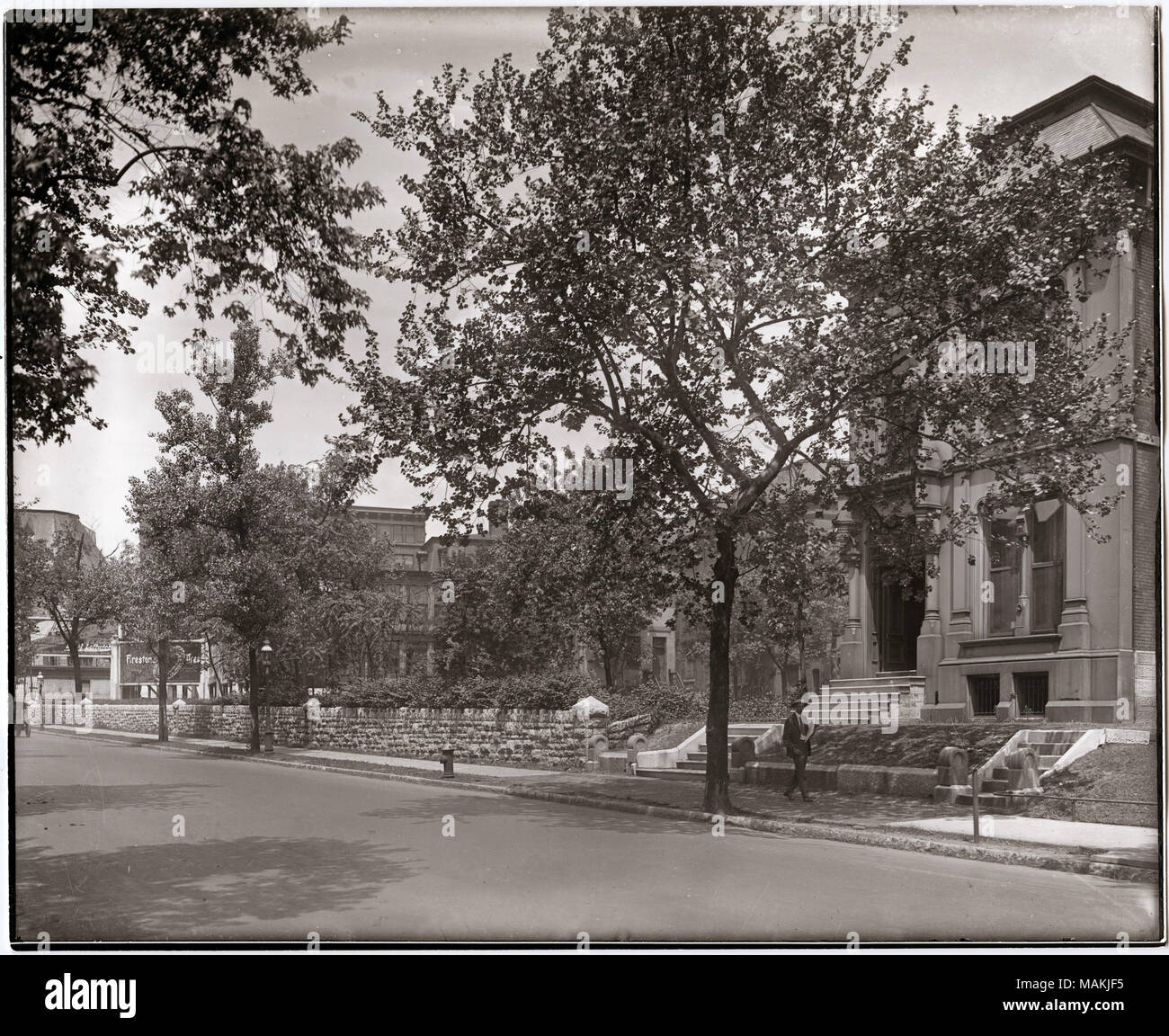 In orizzontale e di fotografia in bianco e nero che mostra Washington vicino al Grand. La strada corre lungo il bordo inferiore della fotografia e un marciapiede può essere visto sul lato opposto della strada. Un basso muro di pietra frontiere molto del marciapiede. Una casa con sculture ornamentali attorno alle finestre e porte è sul lato destro dell'immagine, e un uomo può essere visto passando davanti ad esso sul marciapiede. Altri edifici sono solo parzialmente visibile dietro gli alberi e il fogliame. Una nota manoscritta sul retro della stampa, eventualmente dalla Scuola Suore di Notre Dame, recita: 'Washington vicino al Grand / Stampa dal dottor Sherwi Foto Stock