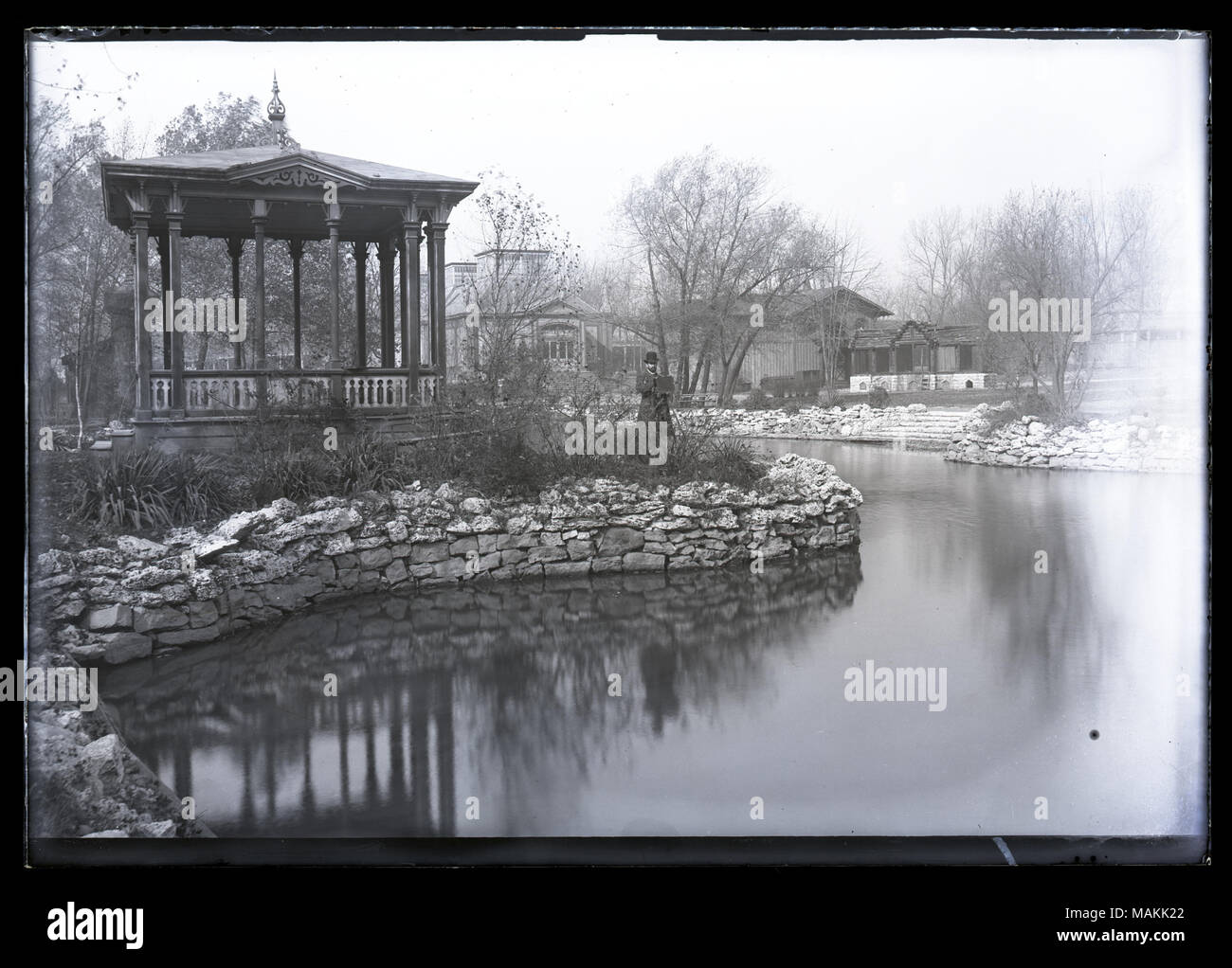 In orizzontale e di fotografia in bianco e nero di un parco in scena con un uomo in piedi accanto a una fotocamera nel centro. Vi è una banda di supporto sul lato sinistro dell'immagine, un lago di avvolgimento attraverso il centro e le case visibili in background. Titolo: William Butler, principal della Yeatman High School, scattare fotografie presso il parco della fiera. . 1894. Foto Stock