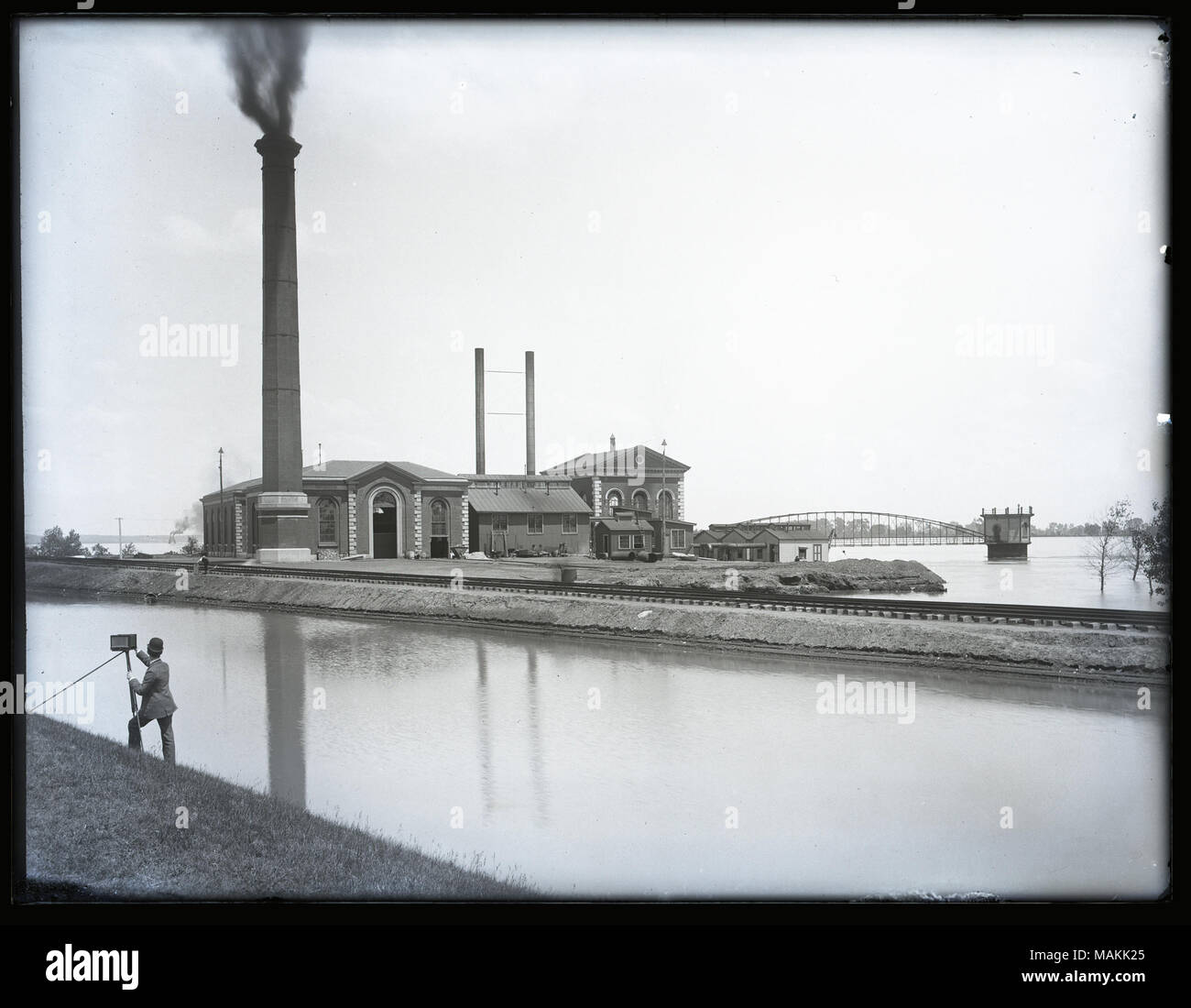 In orizzontale e di fotografia in bianco e nero che mostra un complesso di edifici in mattoni circondata dall'acqua. Vi è un grande fiume con un ponte di distanza e un canale inferiore in primo piano. Nell'angolo in basso a sinistra, un fotografo può essere visto come scattare una foto degli edifici sulla riva opposta del canale. Titolo: catena di rocce acquedotti pubblici Stazione al Riverview Drive. . 1894. Foto Stock
