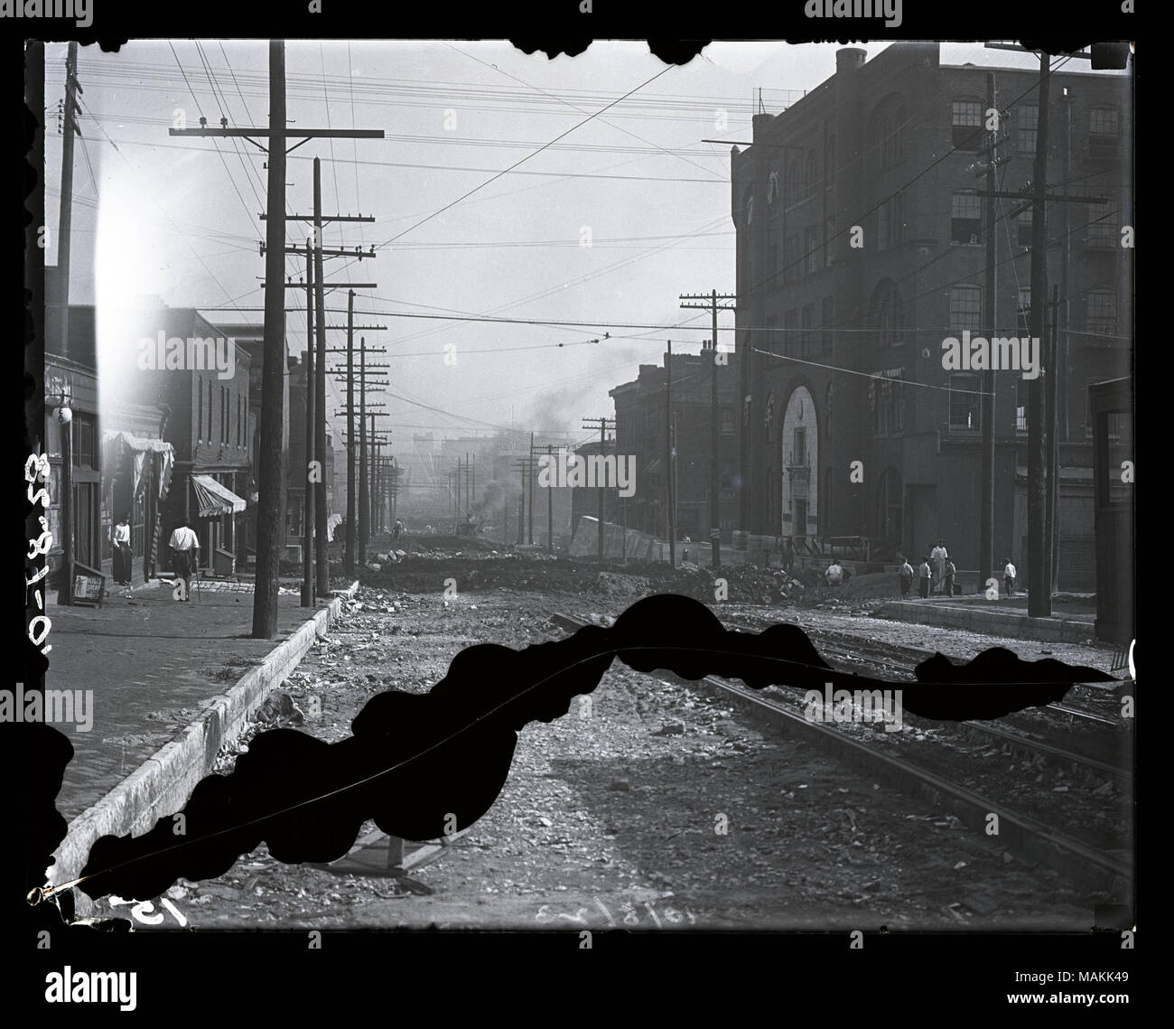 In orizzontale e di fotografia in bianco e nero che mostra un misterioso street scene. La sporcizia street è lacerata, eventualmente per la costruzione di binari del tram. Binari del tram nell'angolo inferiore destro bruscamente fine a metà strada giù per la strada. La costruzione di edifici commerciali può essere visto su entrambi i lati della strada. Ci sono diversi i pedoni a sinistra e un piccolo gruppo di bambini nel piano medio sulla destra. Titolo: Unidentified street scene. . Tra 1890 e 1910. Foto Stock