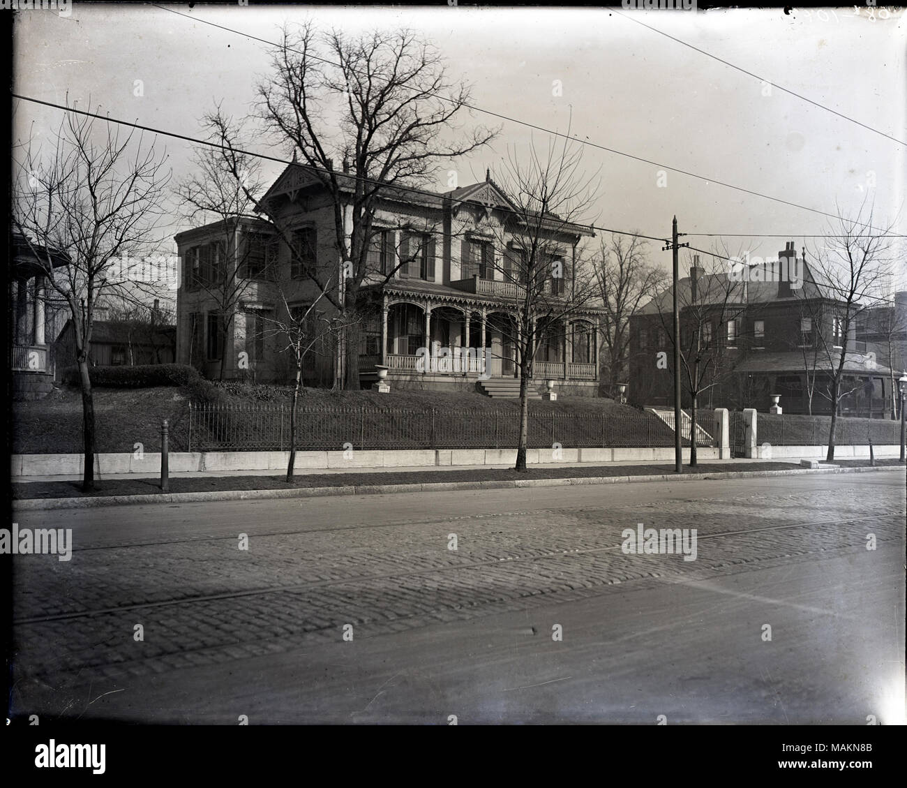 In orizzontale e di fotografia in bianco e nero che mostra due grandi case, probabilmente in Lafayette Avenue-Compton Avenue-Henrietta Street-Louisiana Avenue quartiere. Entrambe le case hanno grandi portici anteriore ed elaborate decorazioni lungo la linea del tetto. Un ferro battuto recinzione confina il marciapiede davanti alle case. Una strada di ciottoli con piste di tram che scorre attraverso il primo piano. La fotografia può essere stato compiuto dalla città di San Luigi a mostrare danni non specificato. Titolo: Grandi Case, probabilmente nel Lafayette-Compton-Henrietta-Louisiana quartiere. . Tra 1900 e 1920. Foto Stock
