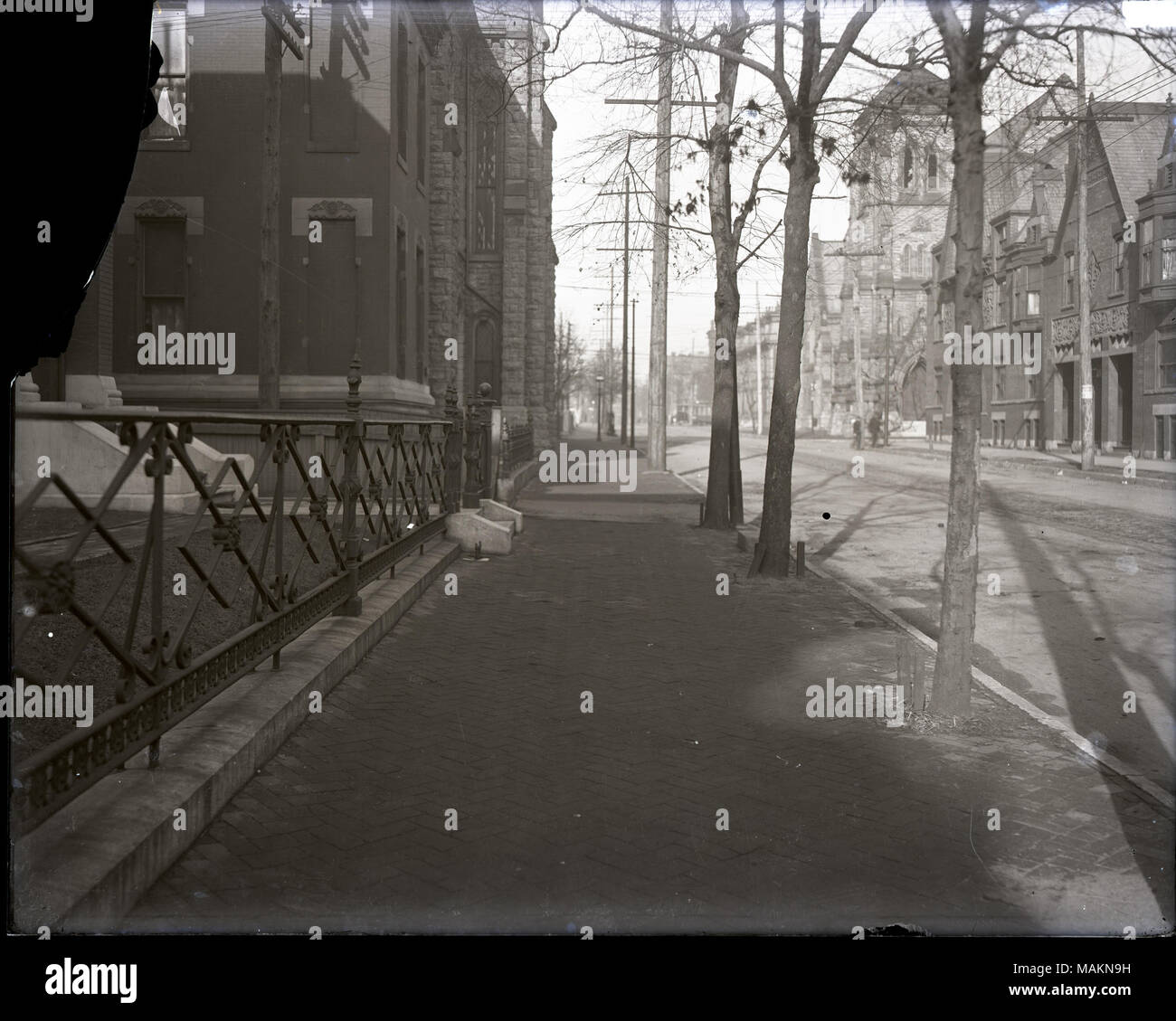 In orizzontale e di fotografia in bianco e nero che mostra una vista guardando verso il marciapiede di mattoni di una strada non identificato. Entrambi i lati della strada sono edifici residenziali accanto a grandi edifici in pietra, eventualmente chiese. In primo piano sulla sinistra vi è il ferro battuto recinto che racchiude un piccolo cortile anteriore. La strada è sulla destra. Titolo: vista guardando giù per una strada non identificato. . Tra 1900 e 1920. Foto Stock